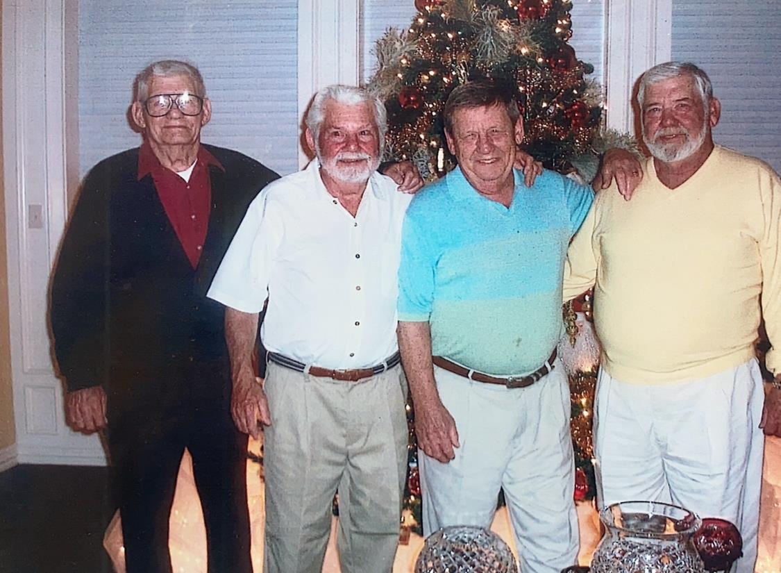 Four older men smile and pose together by a decorated Christmas tree in a warm indoor space.