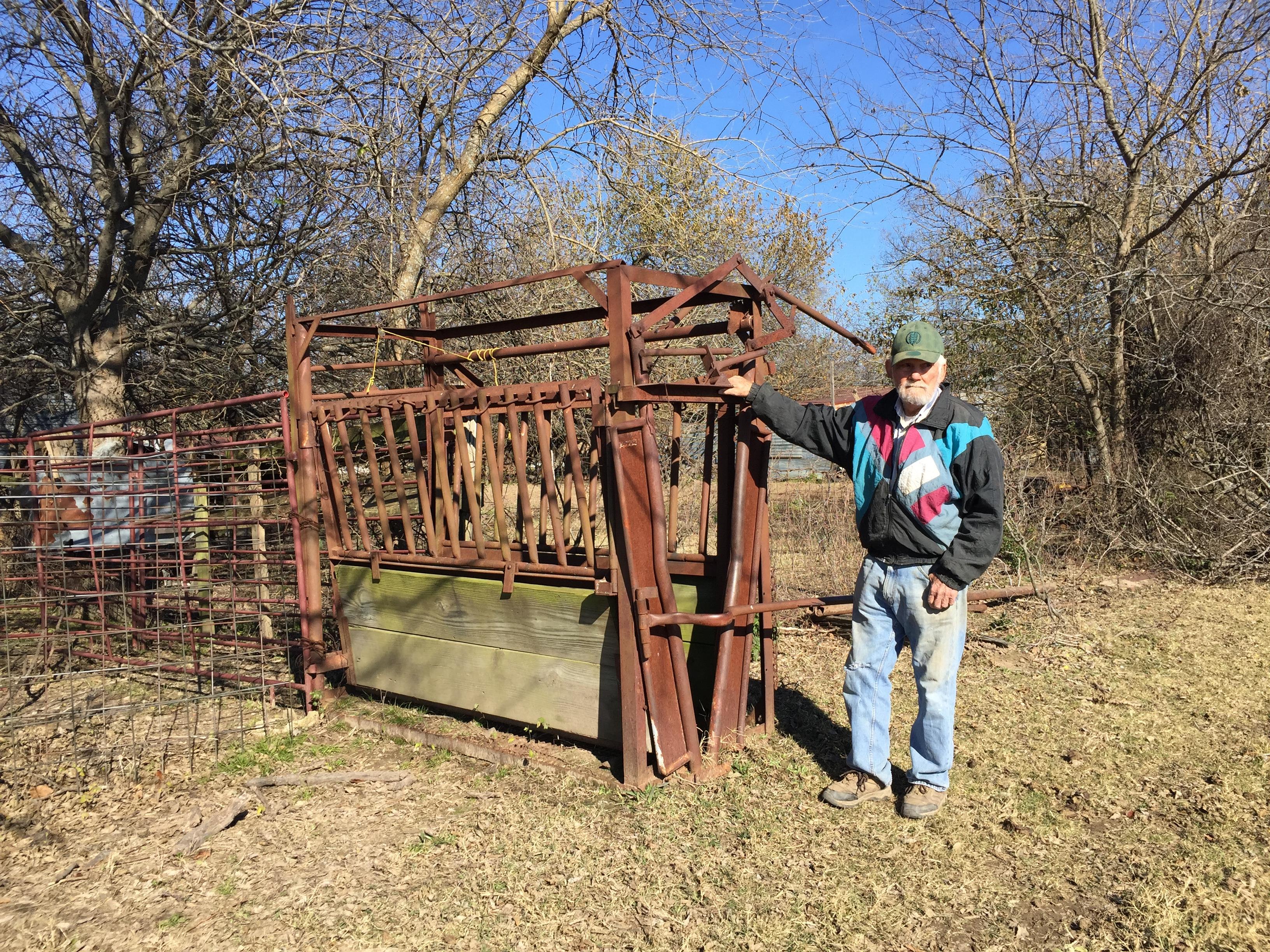 An elderly man leans against rusty farming equipment in a rural area surrounded by trees.