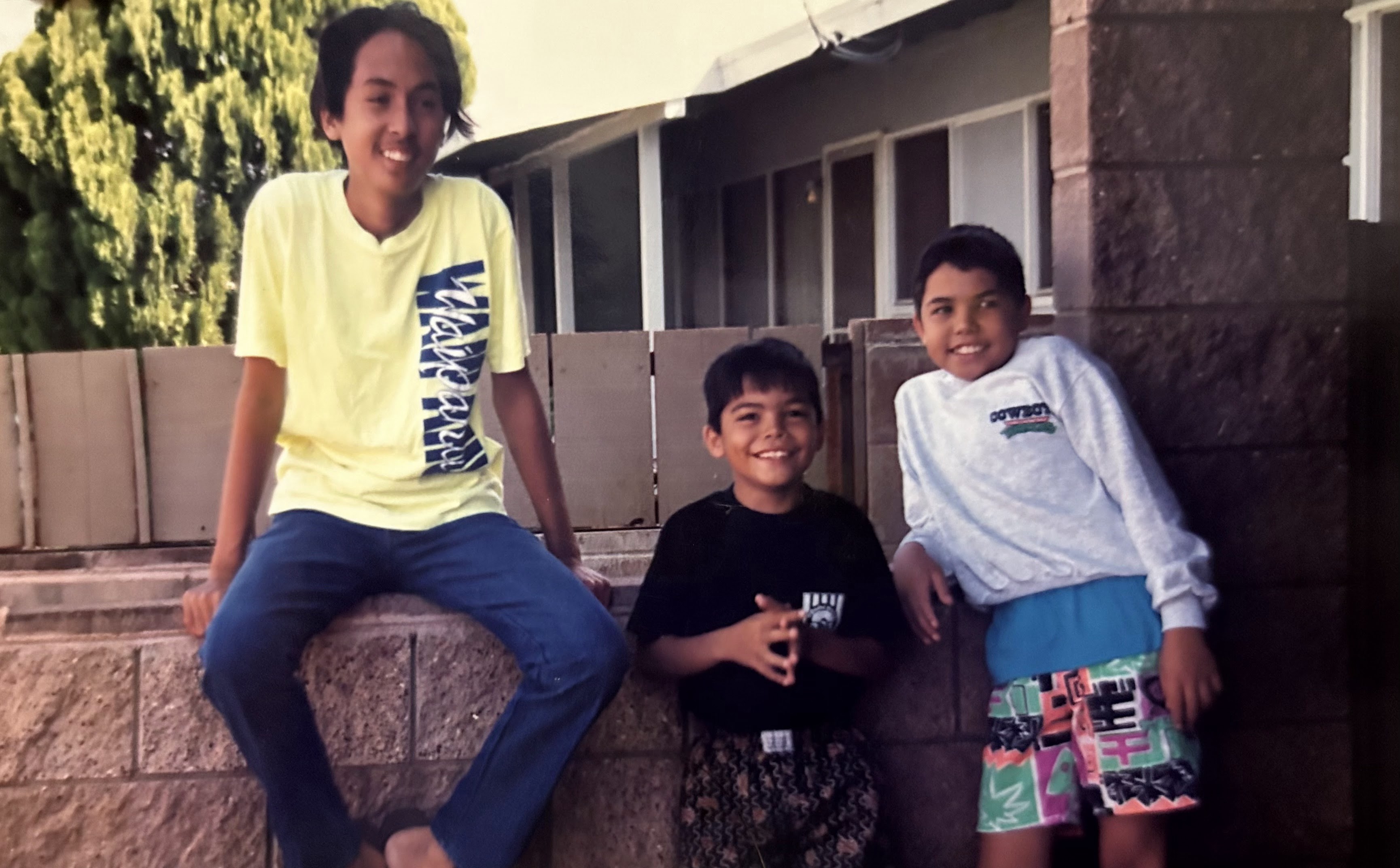 Three boys pose happily outside a house, enjoying a sunny day and smiling for the camera.