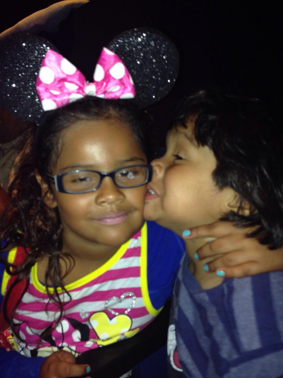Two children share a playful moment, one wearing a bow headband and the other whispering closely.