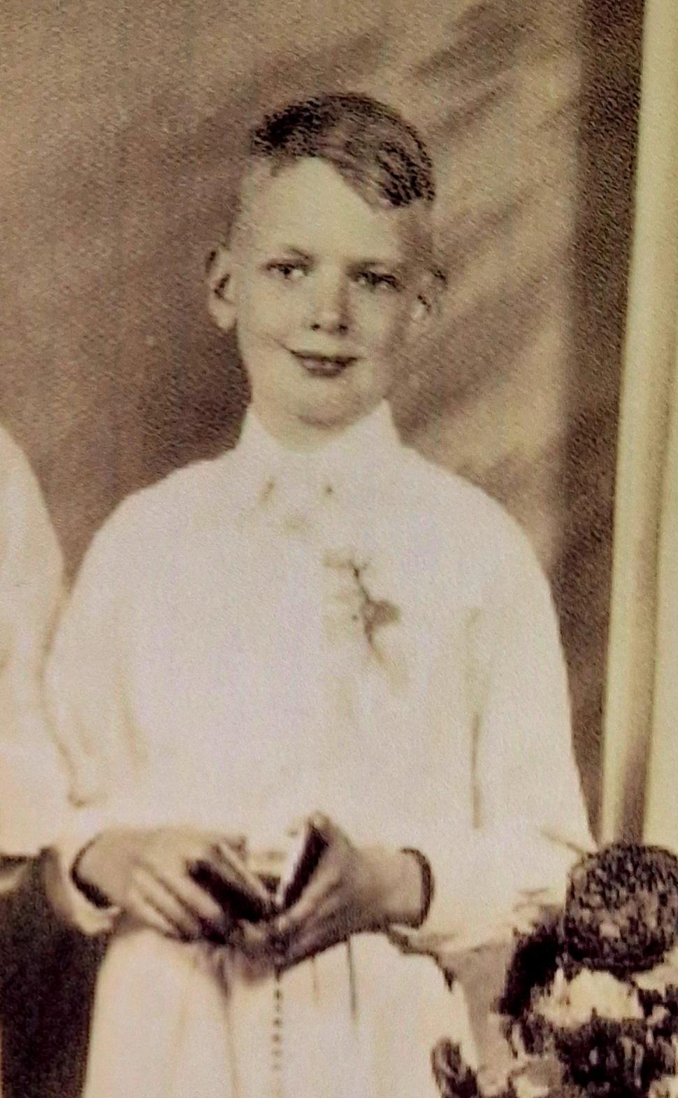 A young boy smiles while holding a gift, dressed in a white outfit during a celebration.