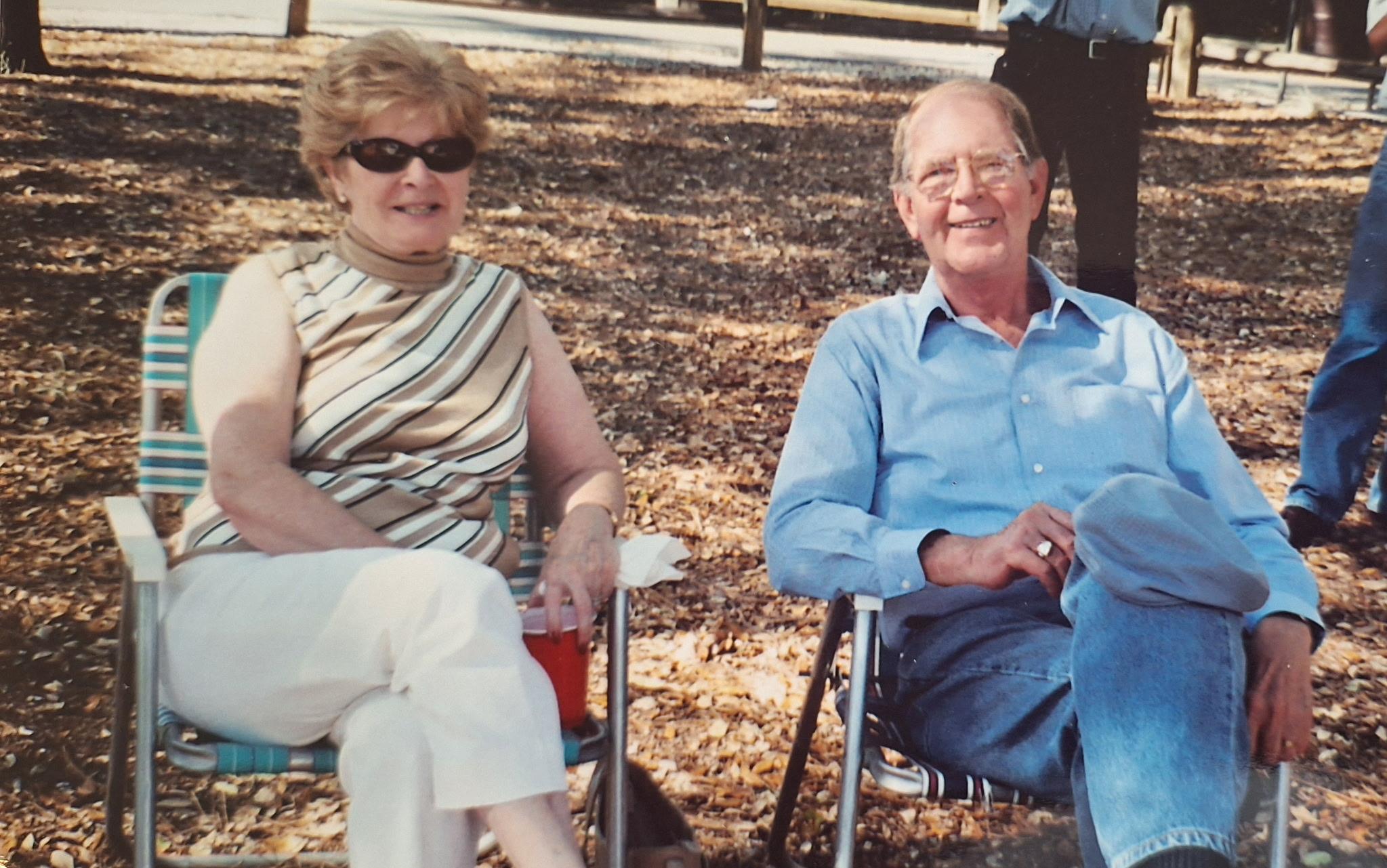 An elderly couple relaxes in folding chairs under sunlight, surrounded by fallen leaves and trees.