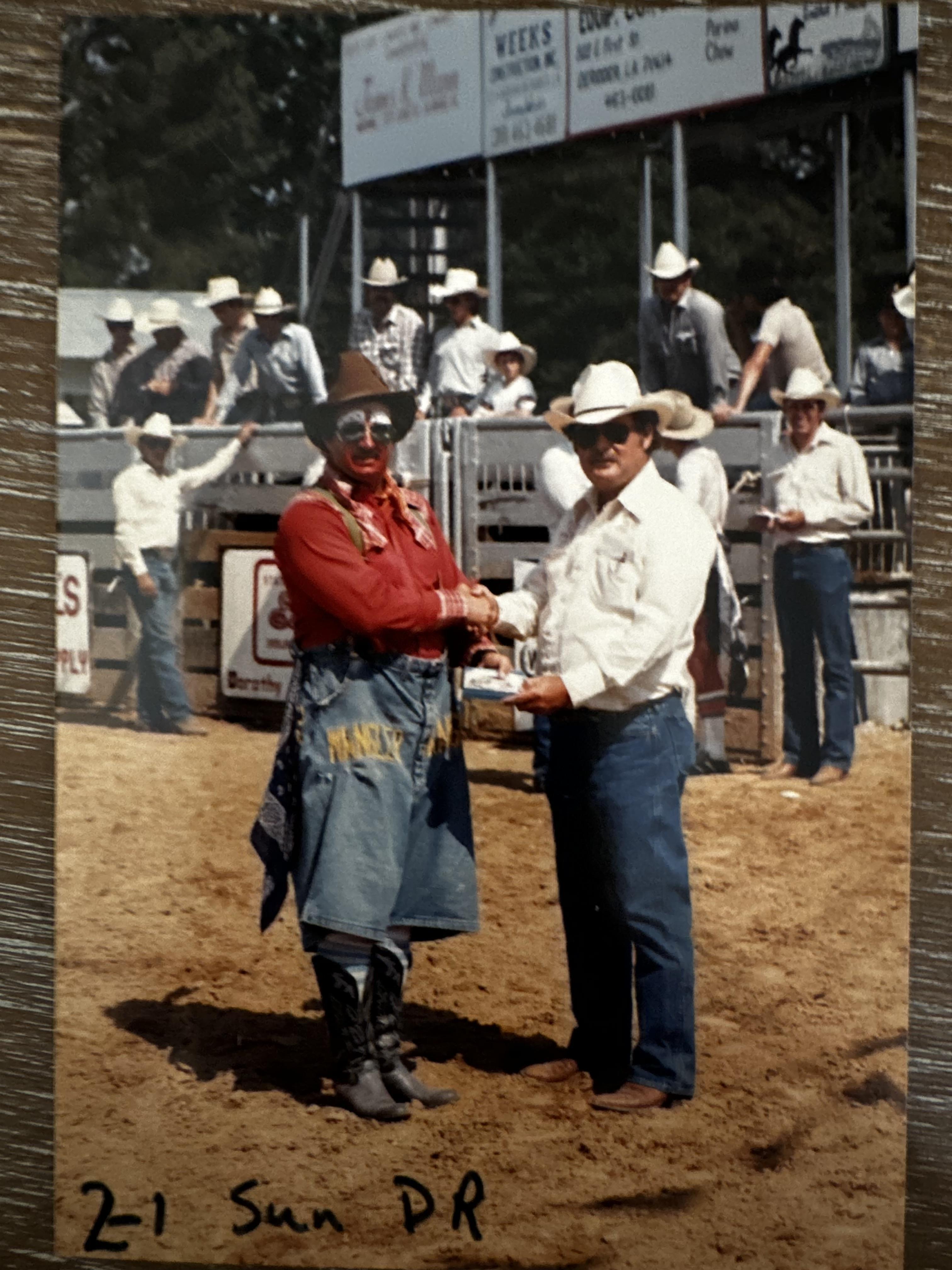 In a lively rodeo setting, a clown and a cowboy shake hands, surrounded by spectators.