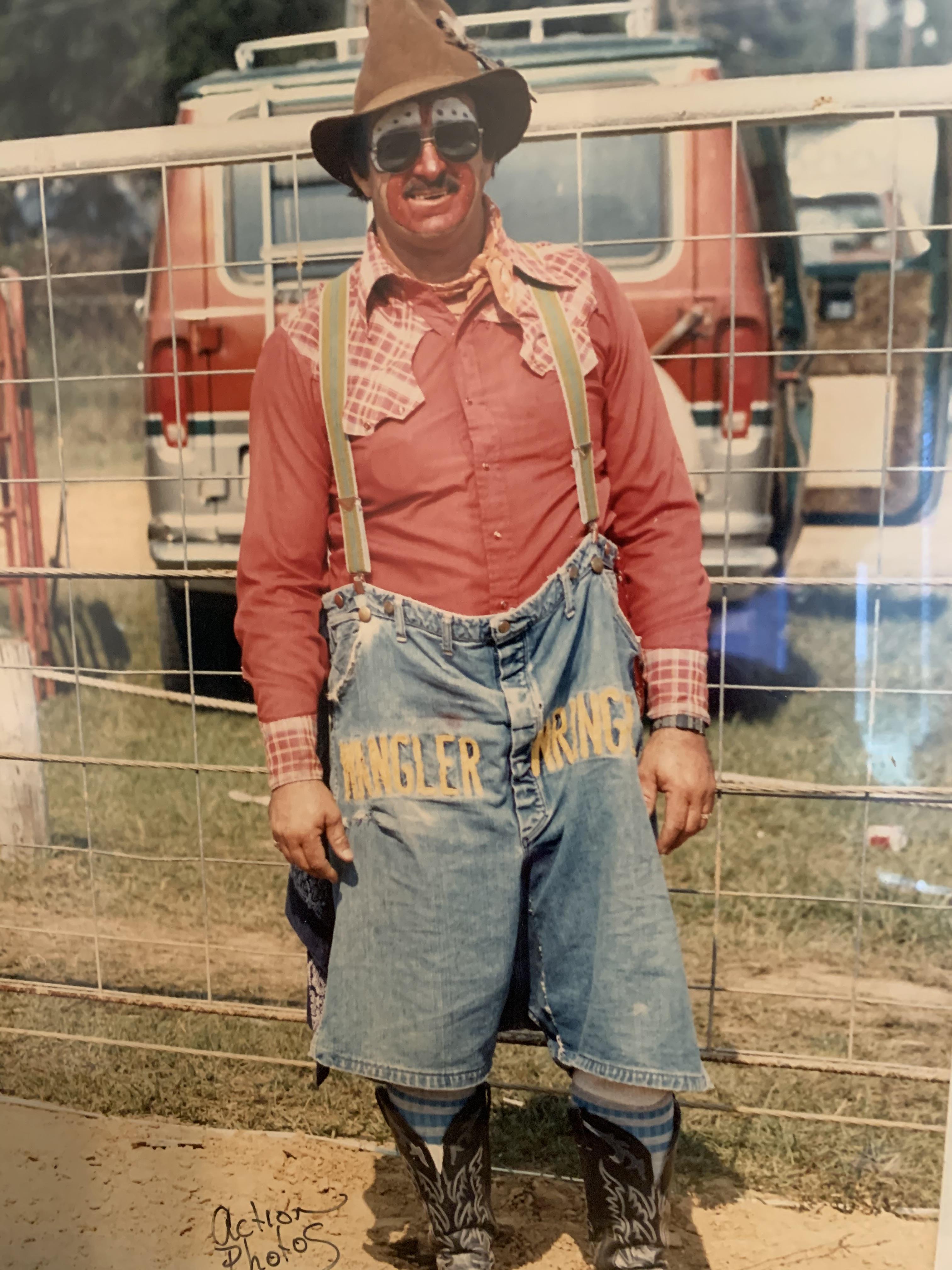 A clown in vibrant clothing stands beside a fence at a lively outdoor gathering.