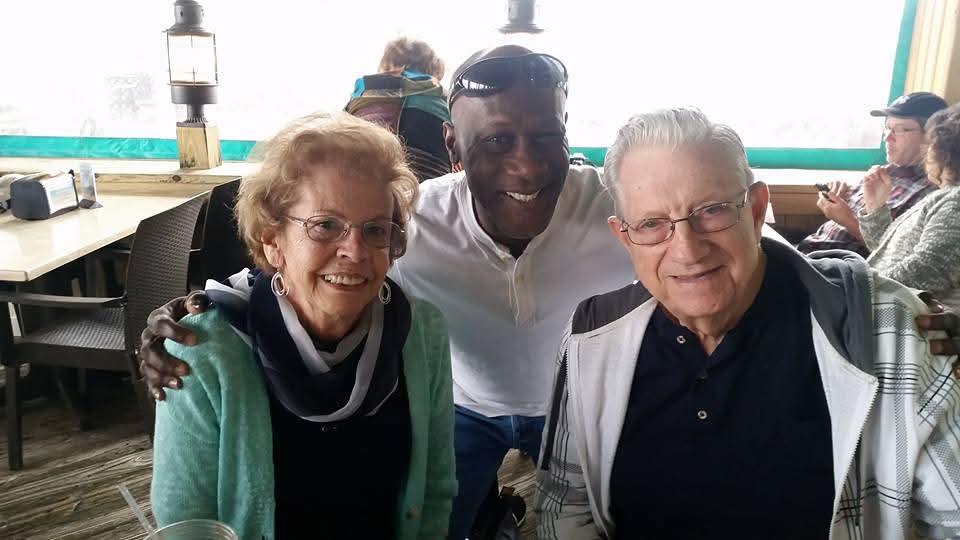 Two seniors share a friendly moment with a smiling man against a lively restaurant backdrop.