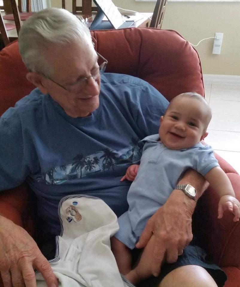 Elderly man smiles warmly while holding a happy baby in a comfortable chair at home.
