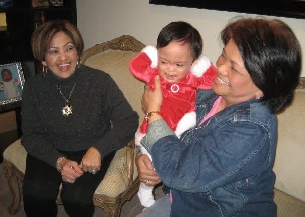 A joyful toddler is playfully embraced by two women during a family gathering at home.