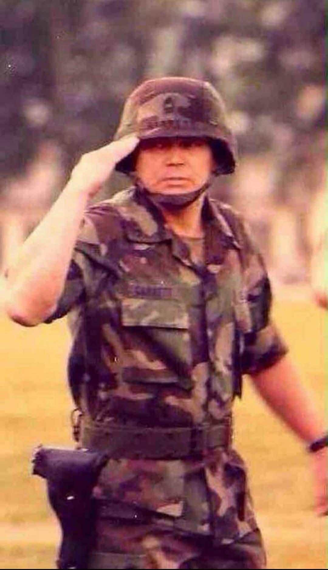 A soldier in camouflage stands at attention, saluting in full military gear.