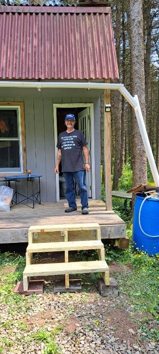 A person in casual attire stands on the steps of a cozy cabin surrounded by tall trees.