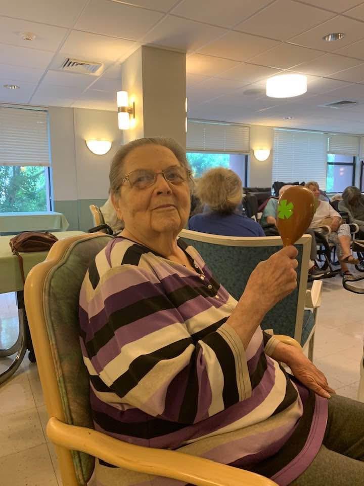Elderly woman smiles while playing maracas in a lively community center filled with guests.