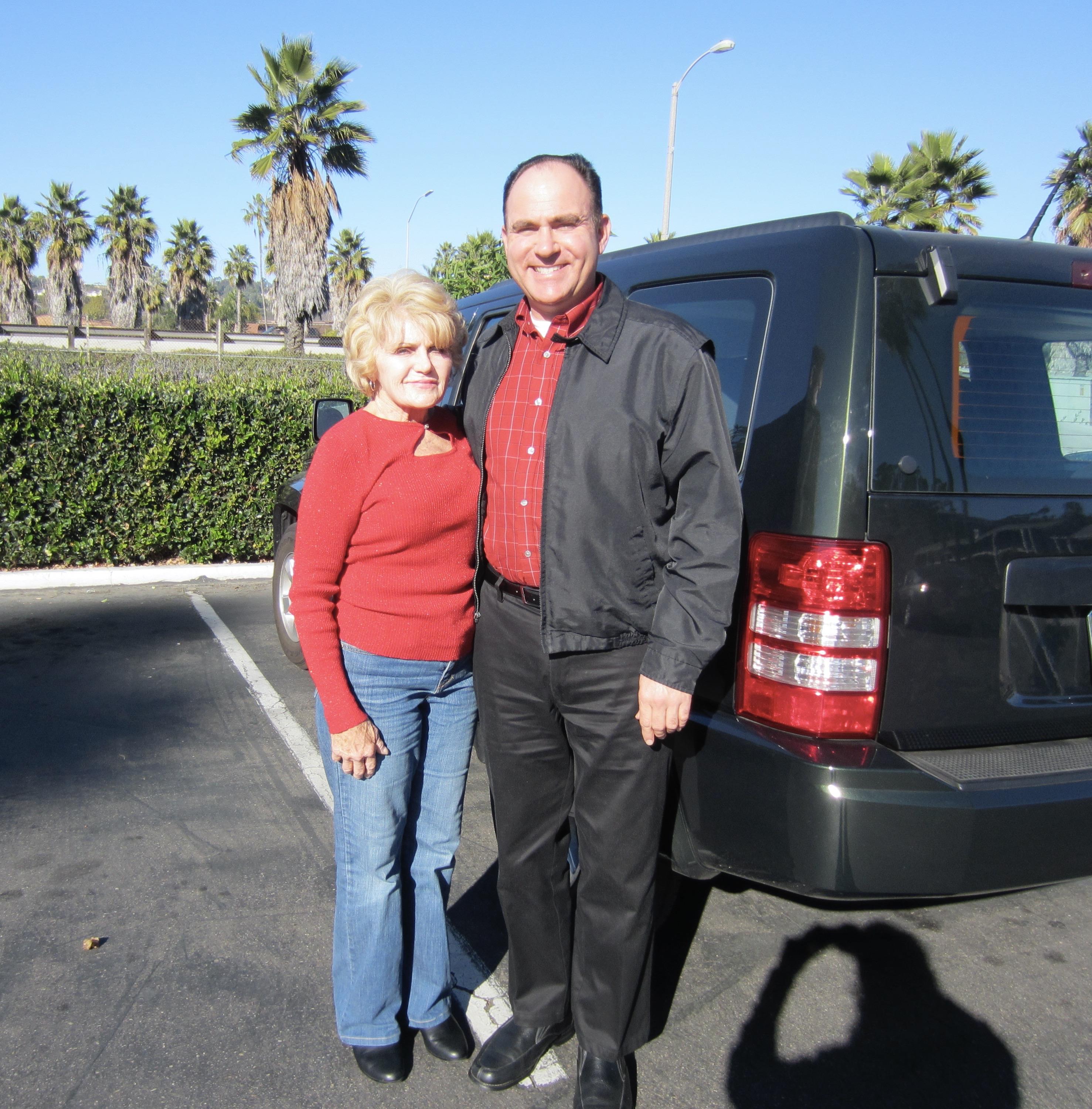 Two smiling individuals stand together near their car under a clear blue sky.