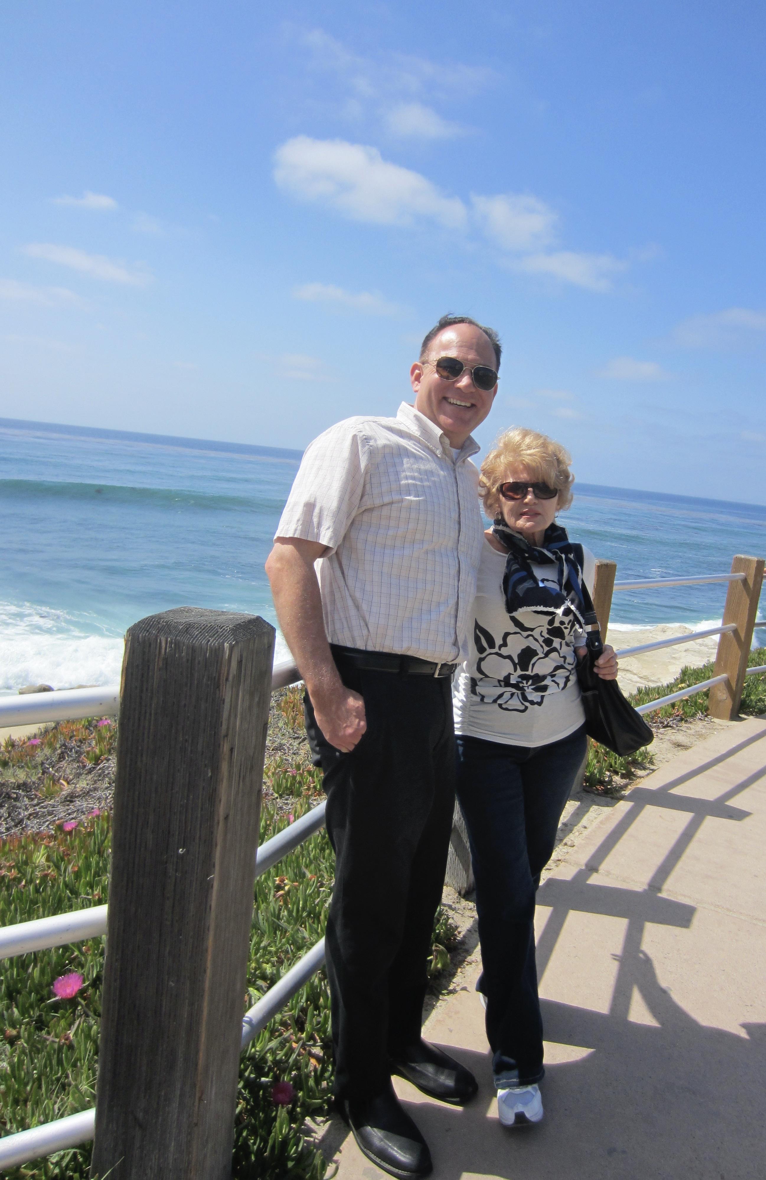 Couple stands together along a coastal pathway, smiling against a backdrop of ocean waves.