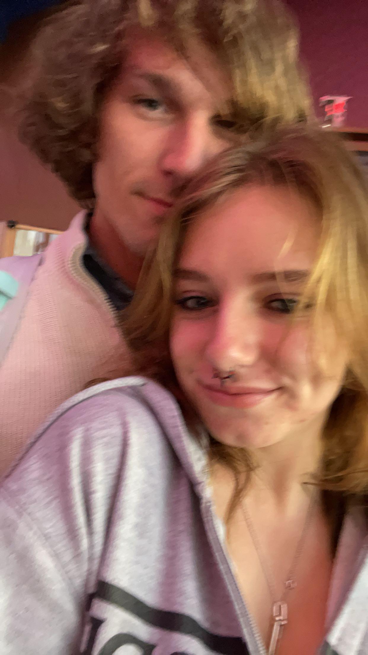 Two individuals stand close together, smiling warmly in a relaxed indoor atmosphere.