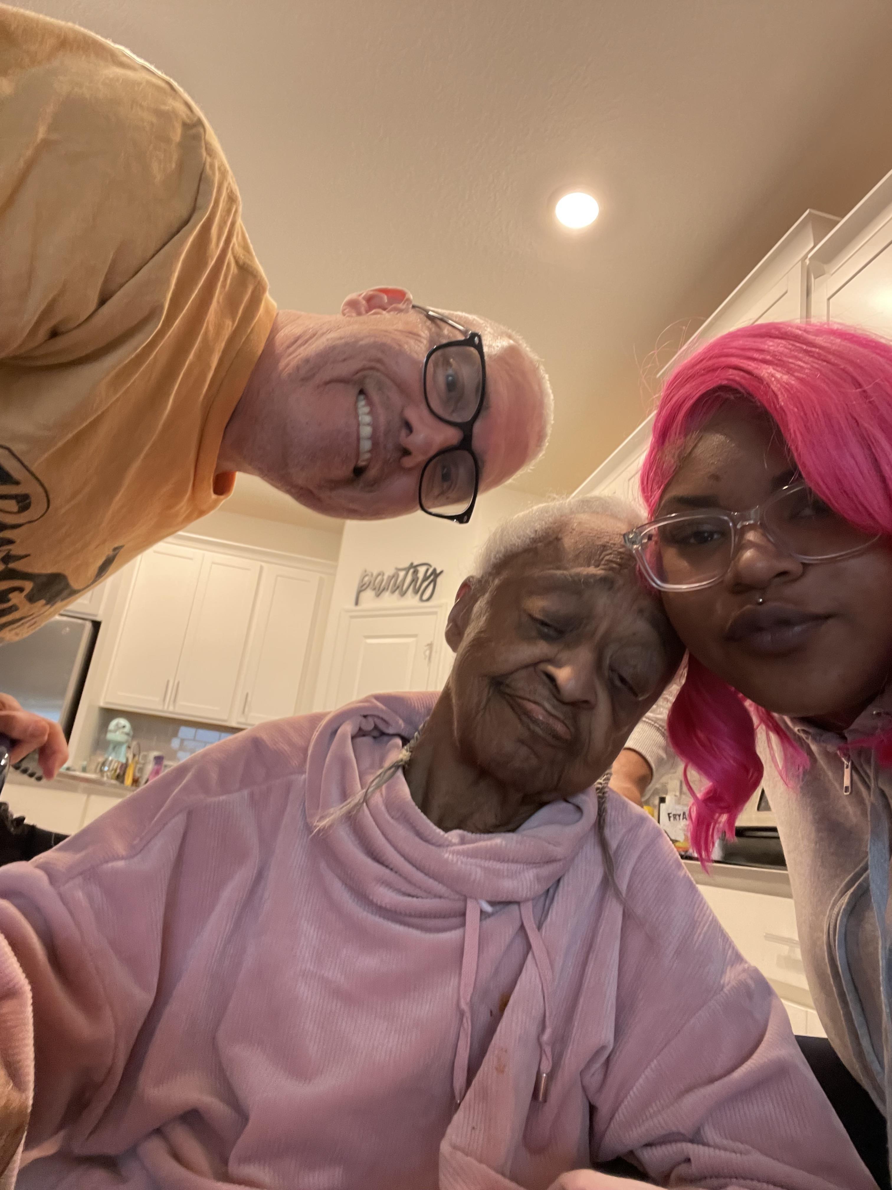 A young woman, an older woman, and a man share a warm moment in a kitchen, smiling together.
