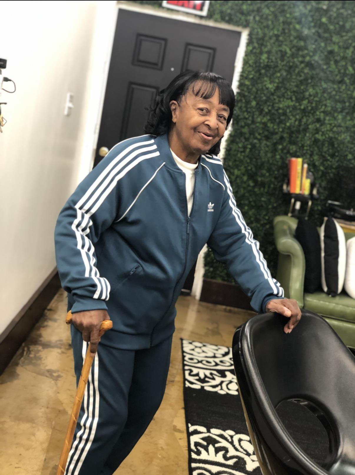 Elderly woman smiles while leaning on a cane, dressed in sporty attire in a cozy interior.