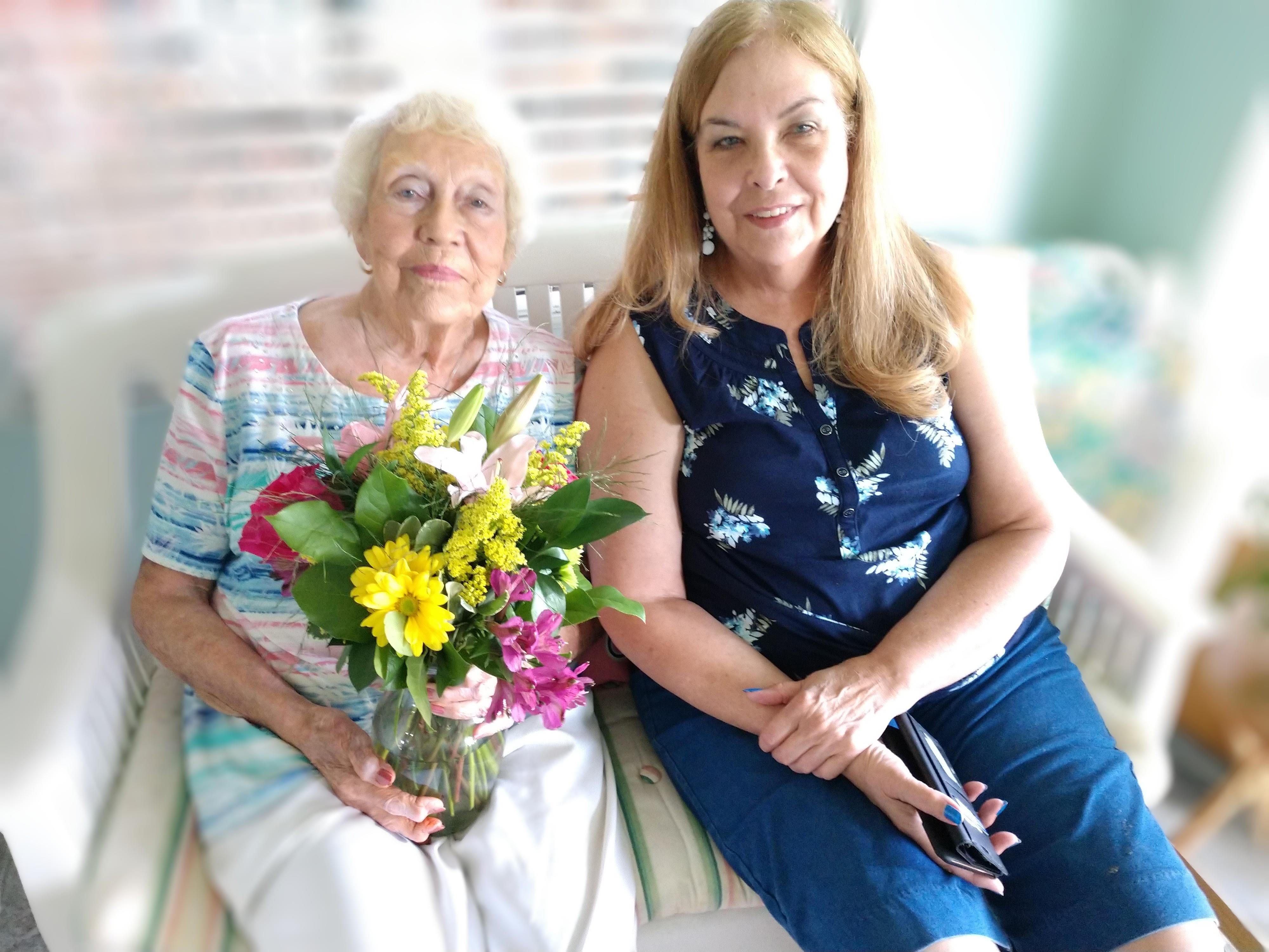 Two women sit together on a bench, surrounded by light decor, holding a vibrant flower bouquet.