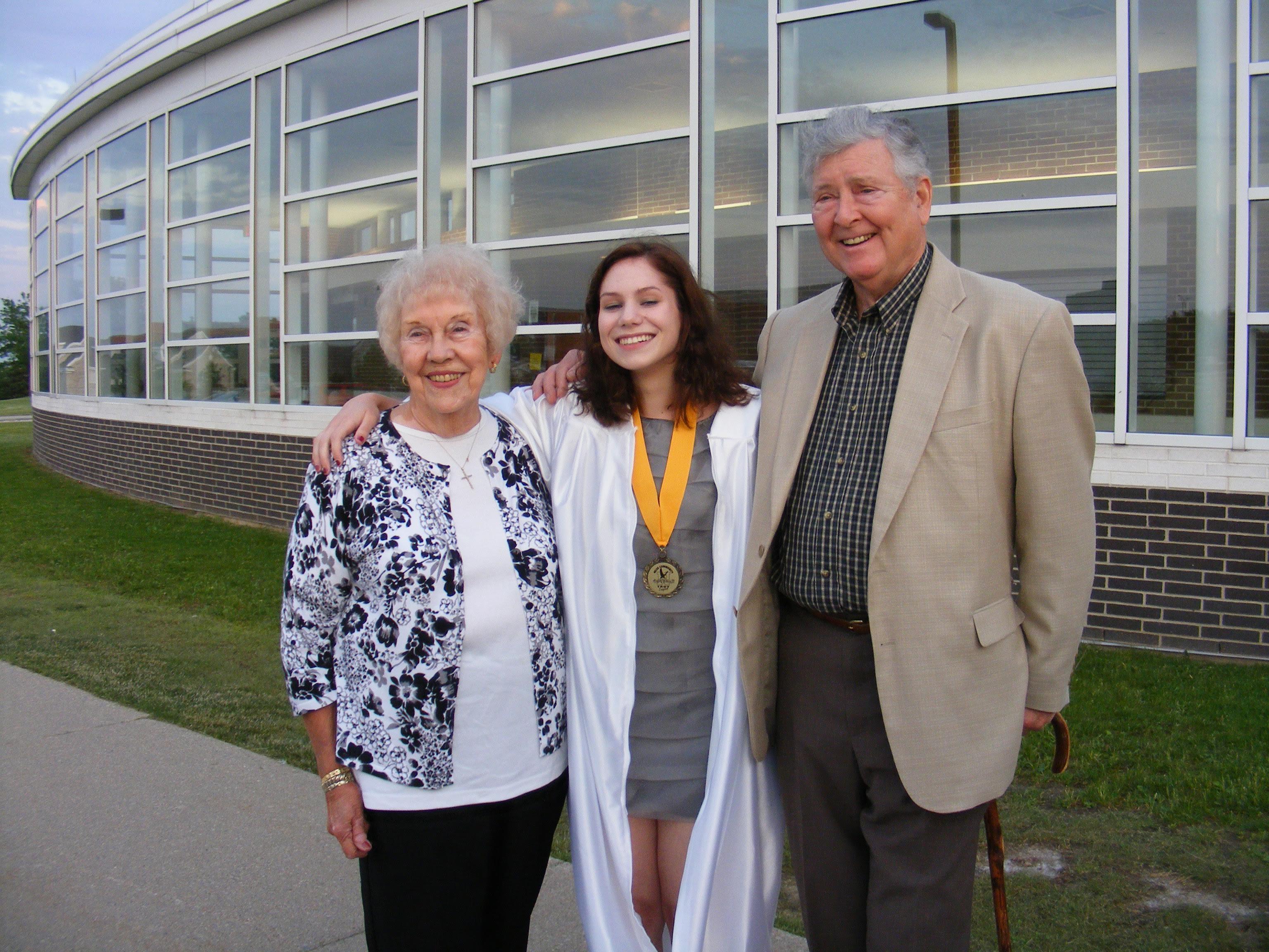 Young woman in graduation attire poses happily with her grandparents outside school.
