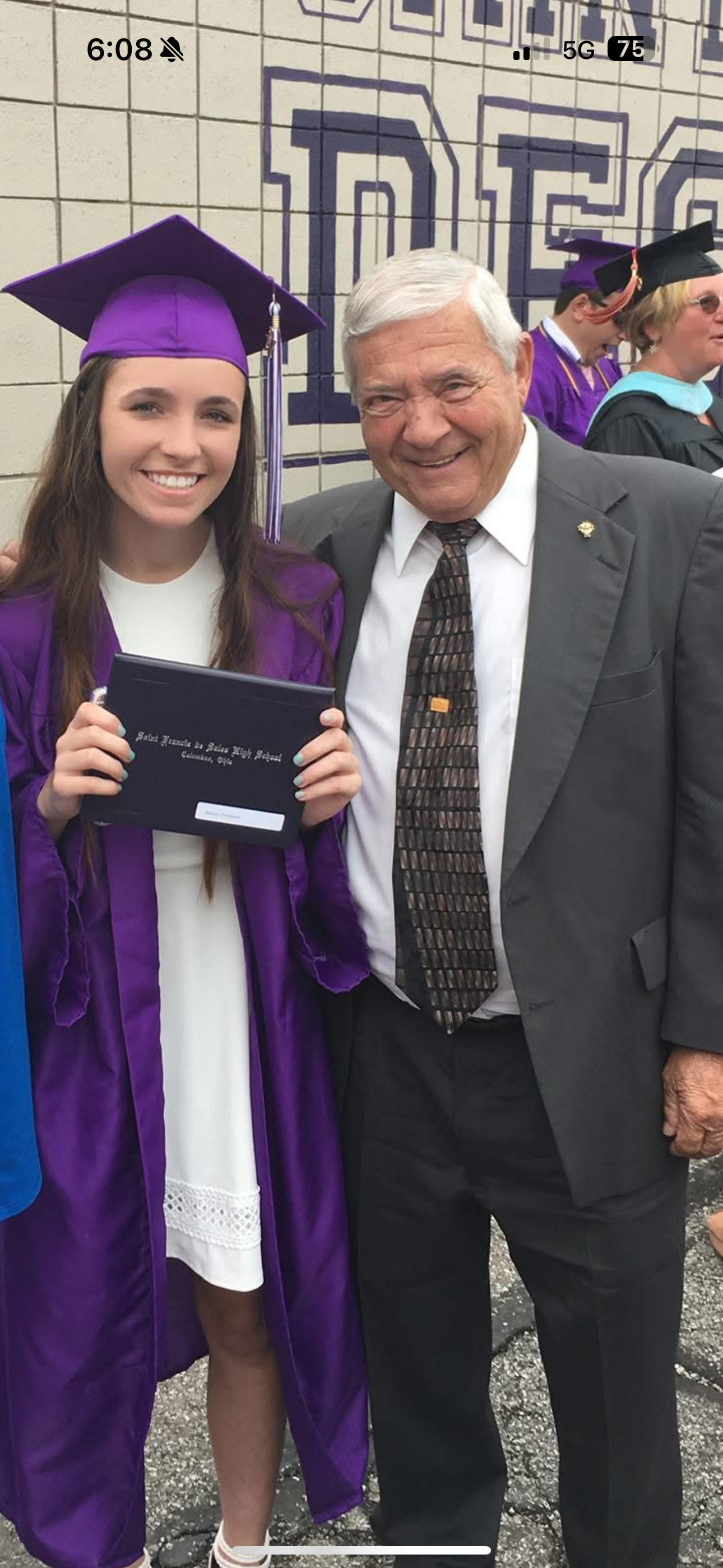 Graduate in cap and gown smiles proudly holding diploma alongside a relative at the ceremony.