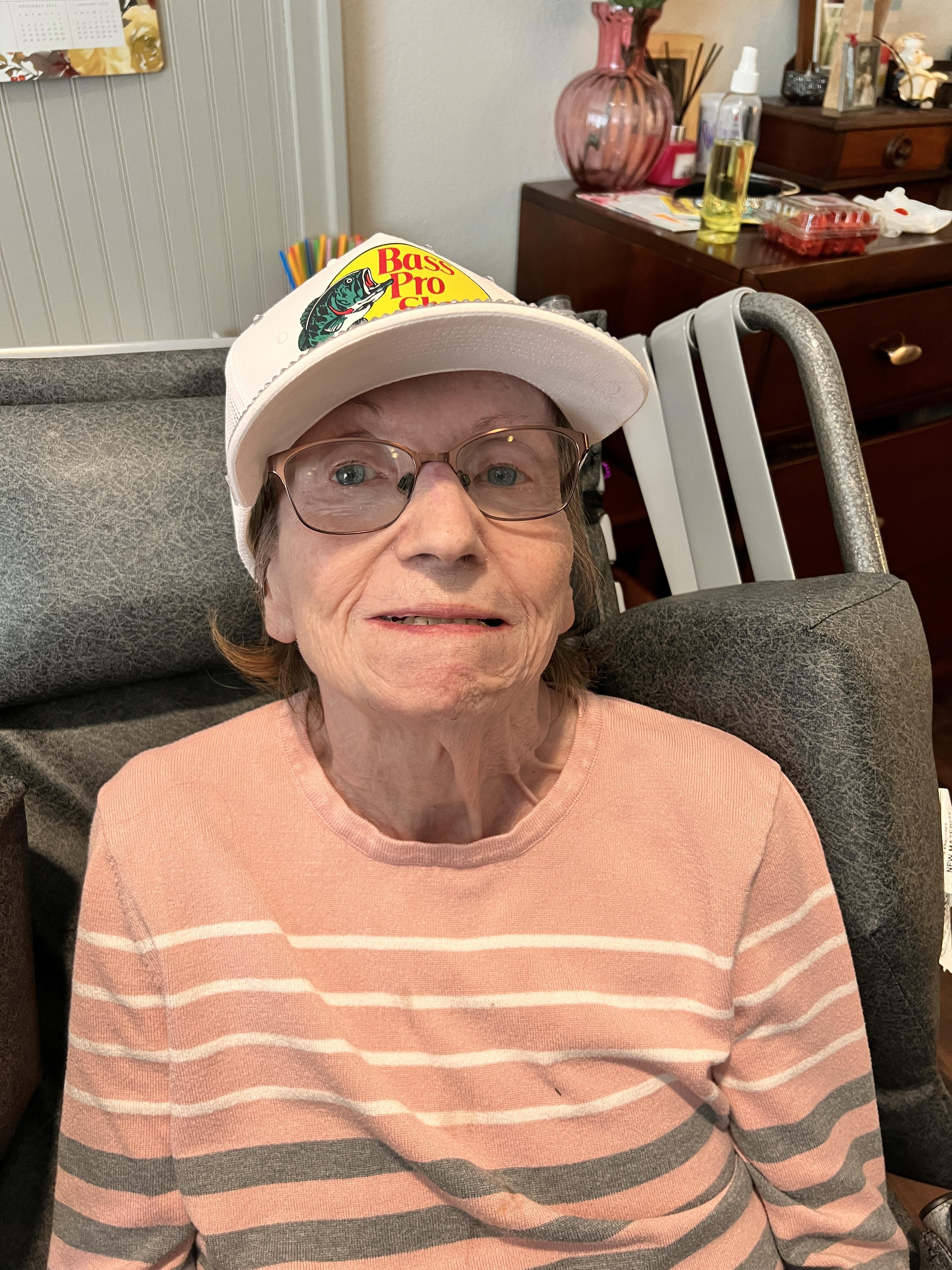 An elderly woman with glasses and a white hat smiles warmly while sitting comfortably indoors.