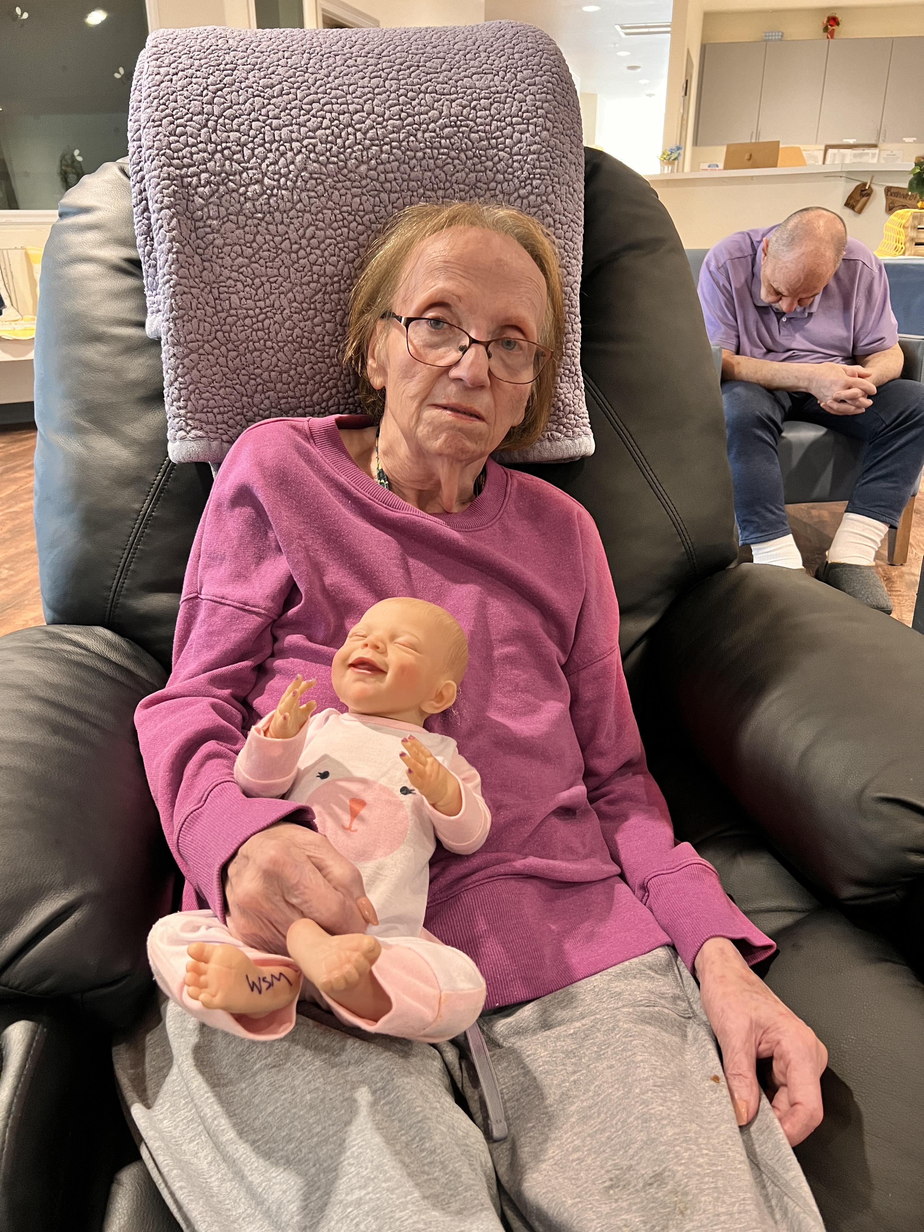 An elderly woman sits comfortably in a chair, cradling a doll made to look like a baby.