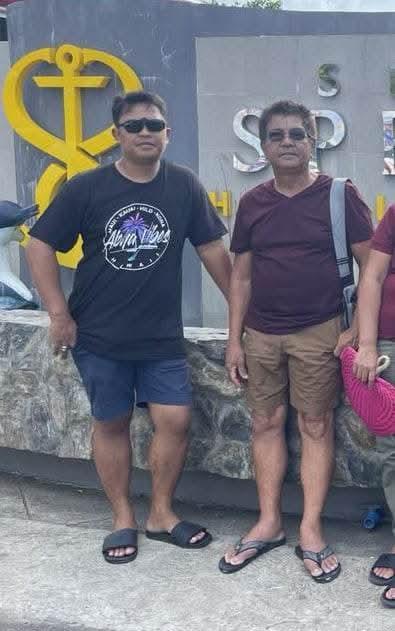Two friends stand together smiling near a marina, enjoying a sunny afternoon by the water.
