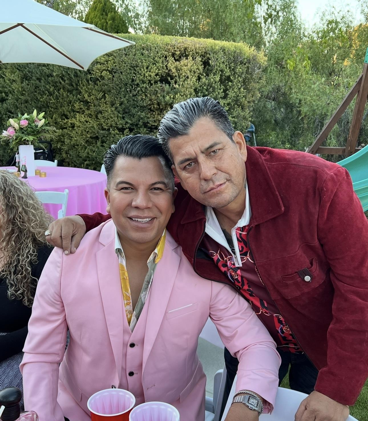 Two men smile warmly at the camera while enjoying a social gathering outdoors in a garden.
