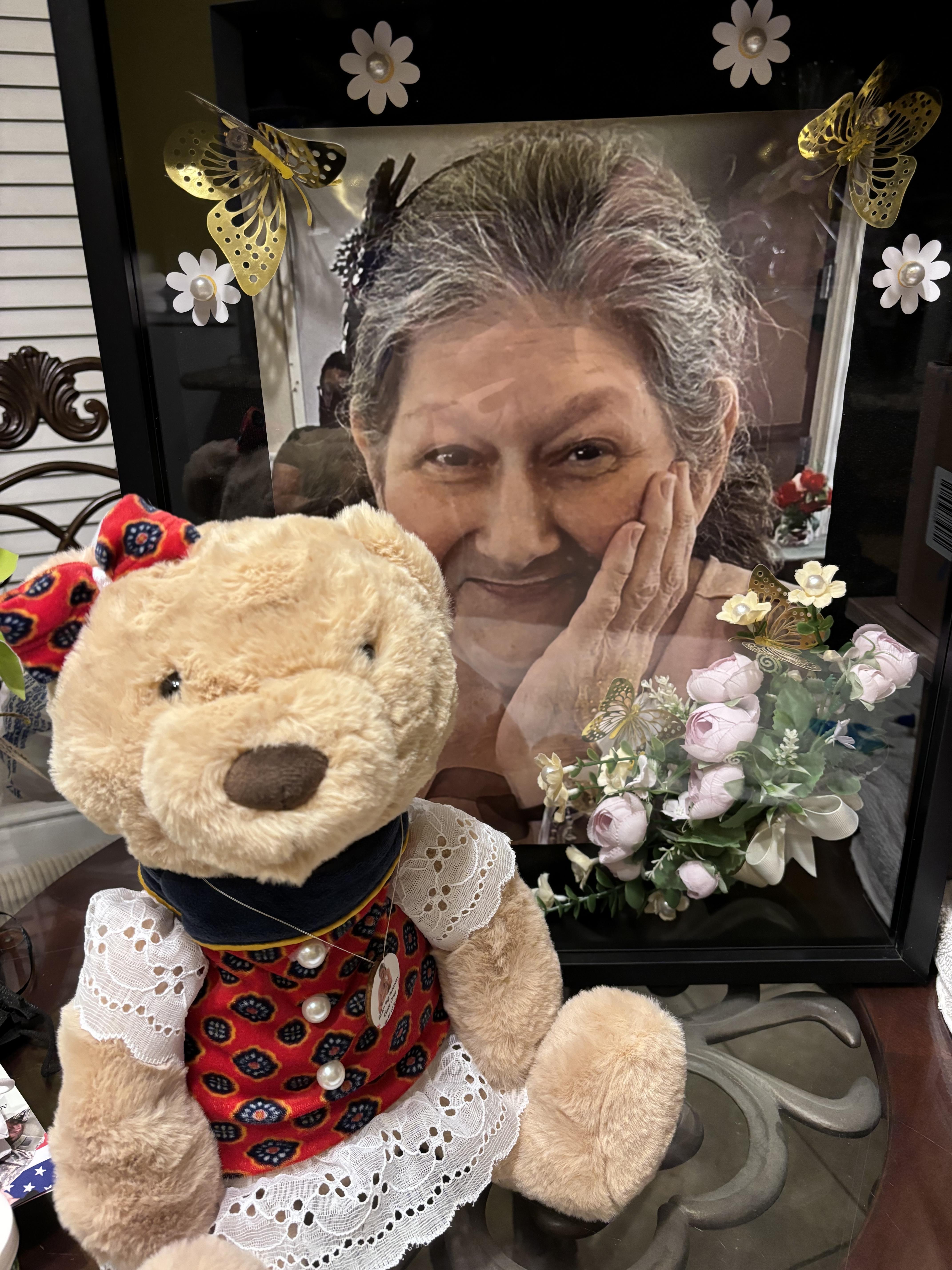 A teddy bear dressed in cultural clothing sits beside a portrait of a woman with a warm smile.