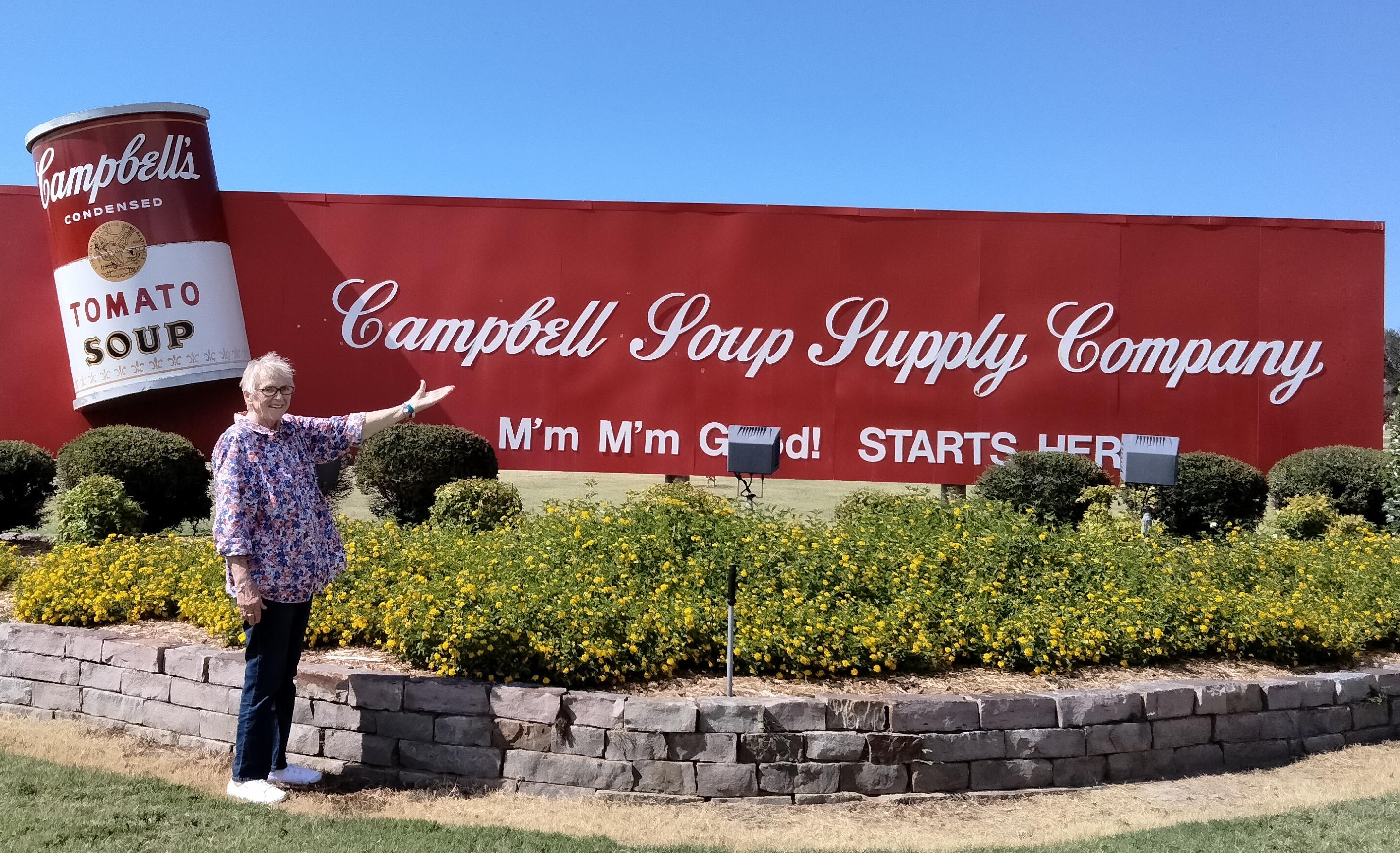 Individual points towards the large Campbell Soup sign surrounded by greenery and flowers.