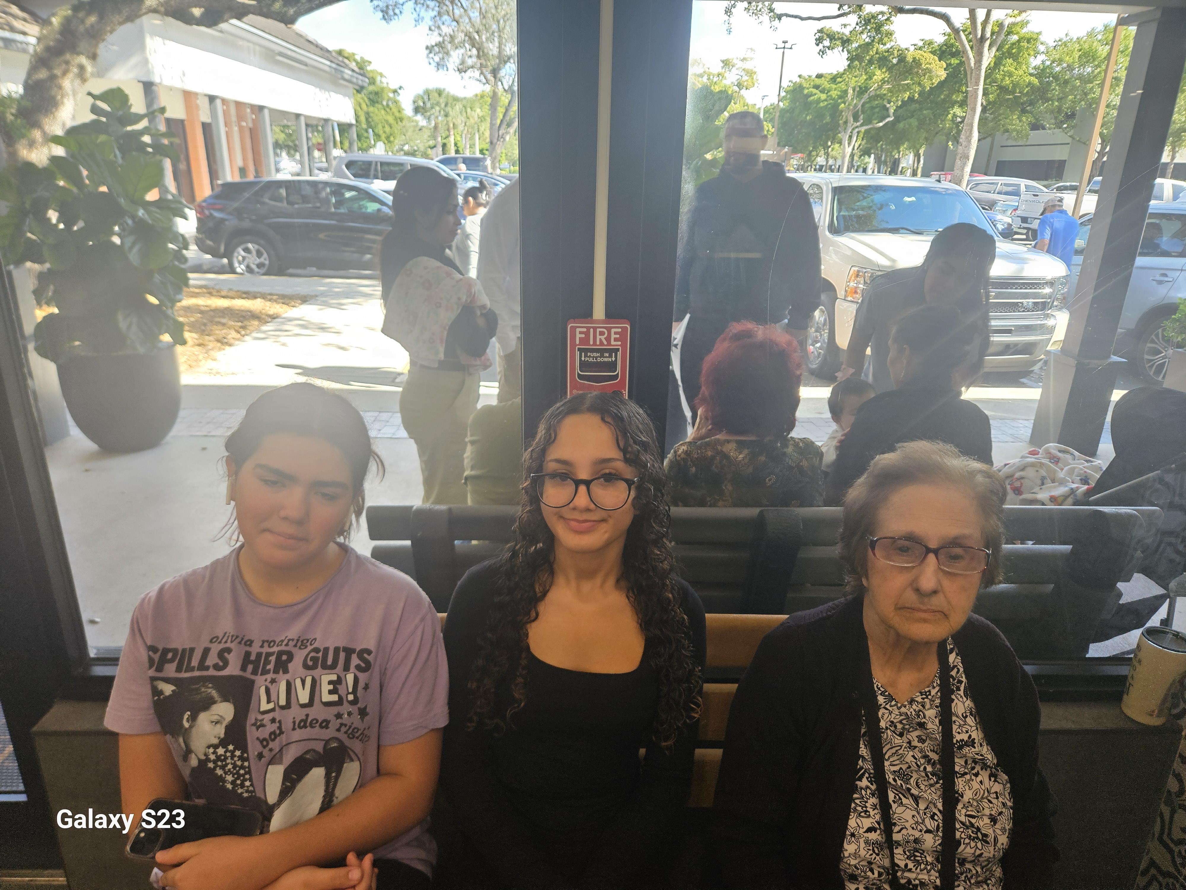 Three women sit in a waiting area while various people are visible outside in the background.