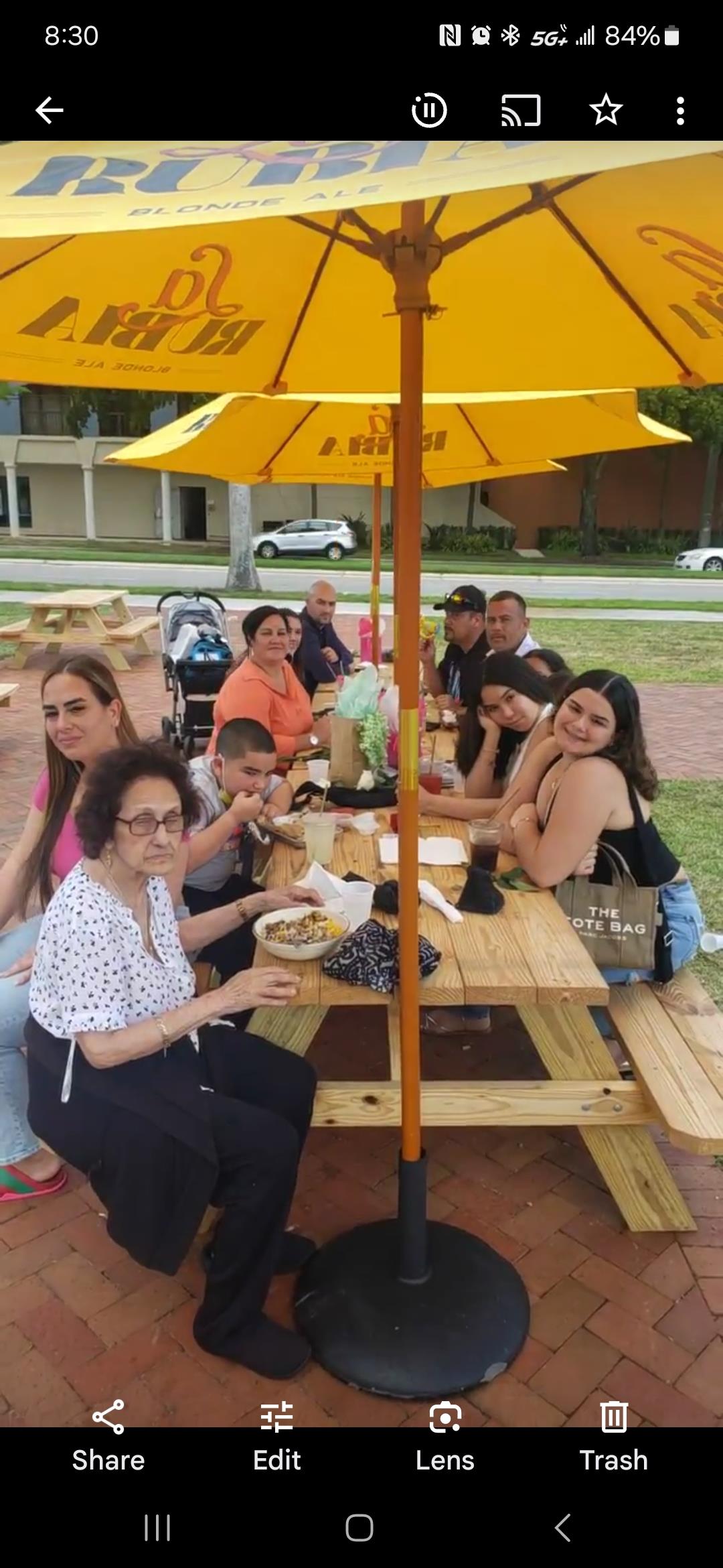 Group of people enjoying food and drinks together at a park picnic table under yellow umbrellas.