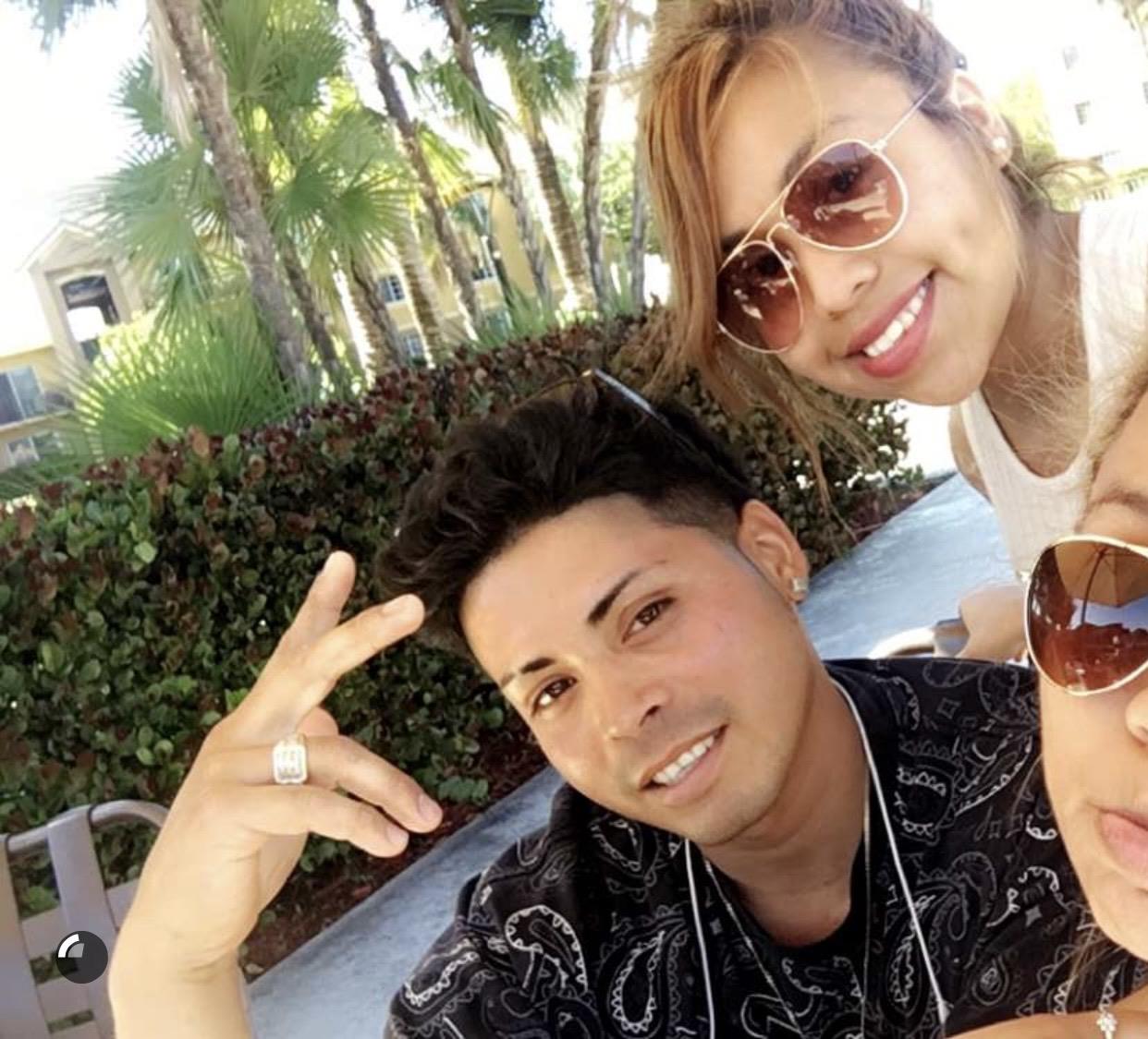 Three friends relax outdoors, smiling and posing together in bright sunshine surrounded by greenery.