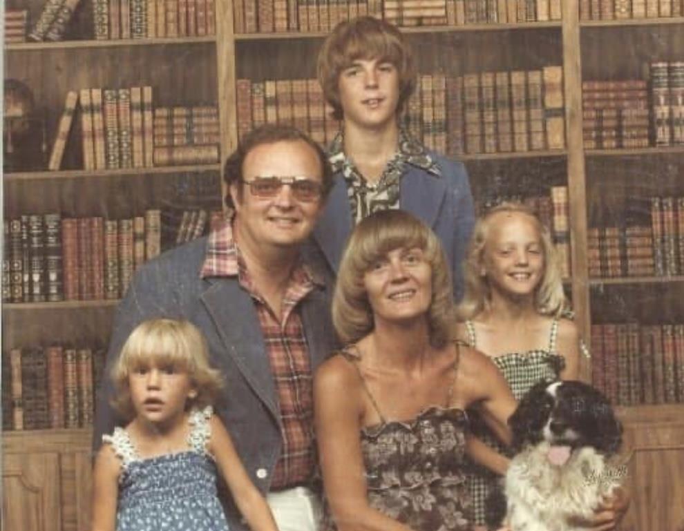 Smiling family poses together in a vintage library, surrounded by books and their pet dog.