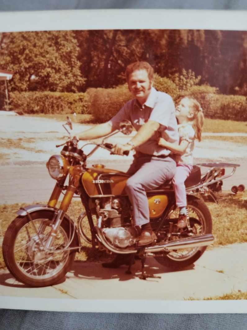 A man and a young girl enjoy a joyful ride on a vintage motorcycle in a vibrant yard.