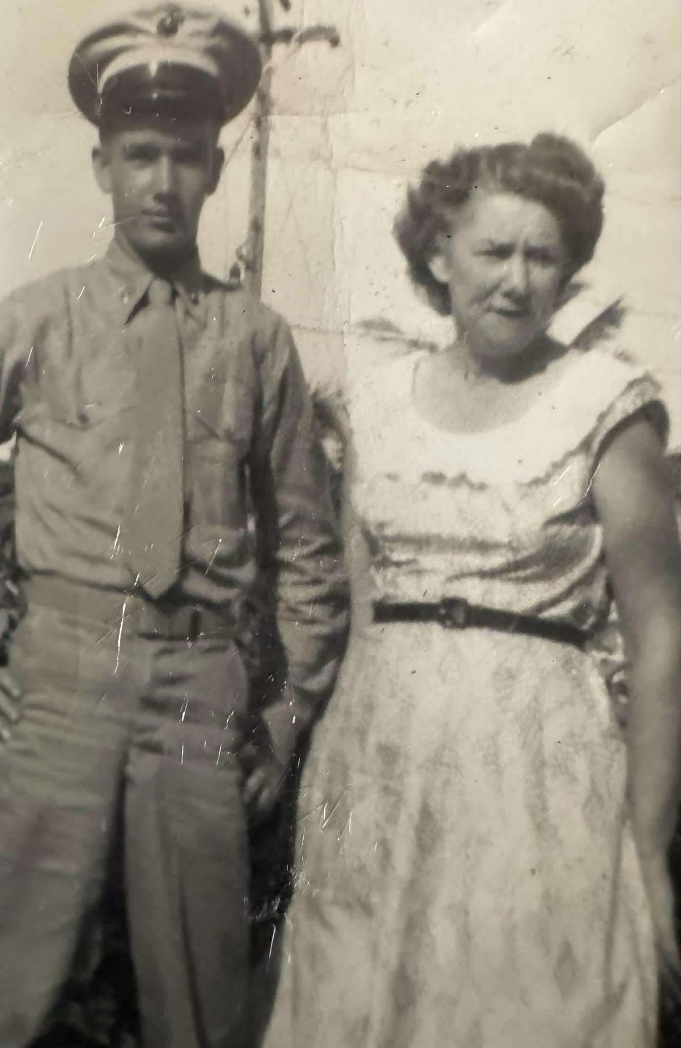 A soldier in military uniform poses with a woman in a light dress outdoors.