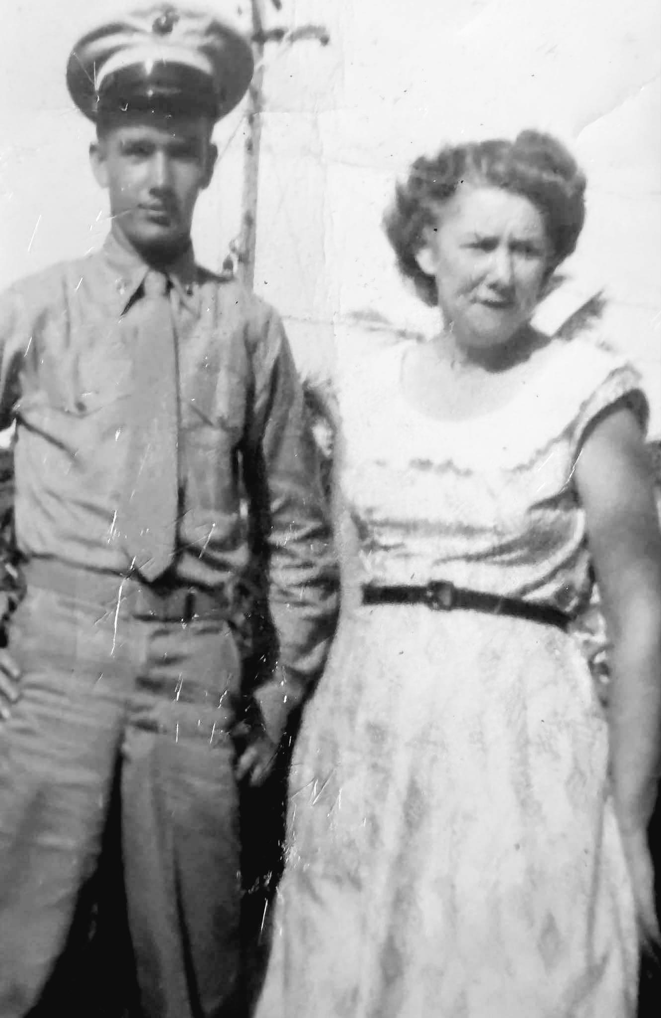 A young soldier stands next to a woman in a flowing dress in a sunny outdoor setting from the past.
