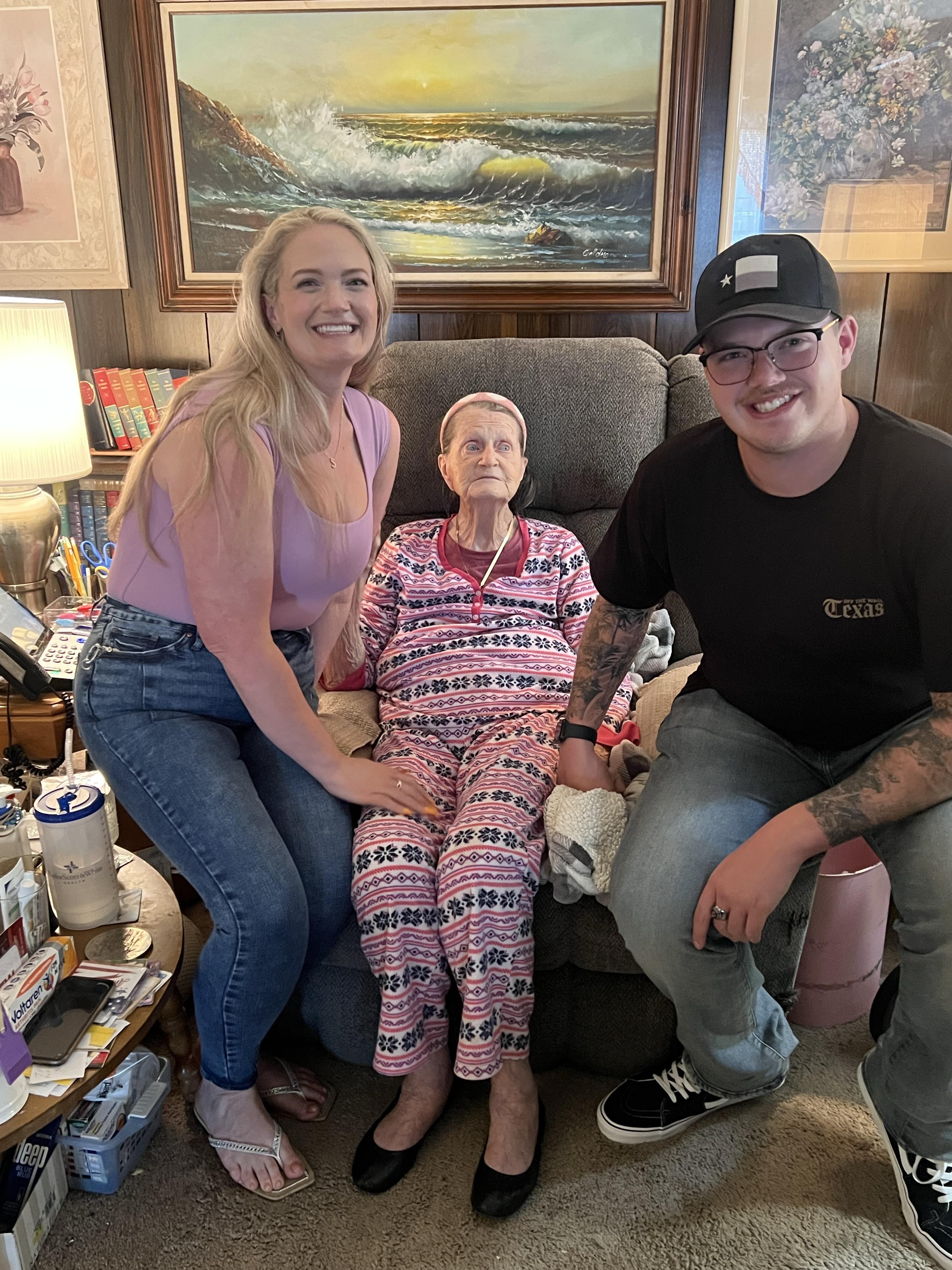 Two younger adults smile while posing with their grandmother in her living room, filled with warmth.