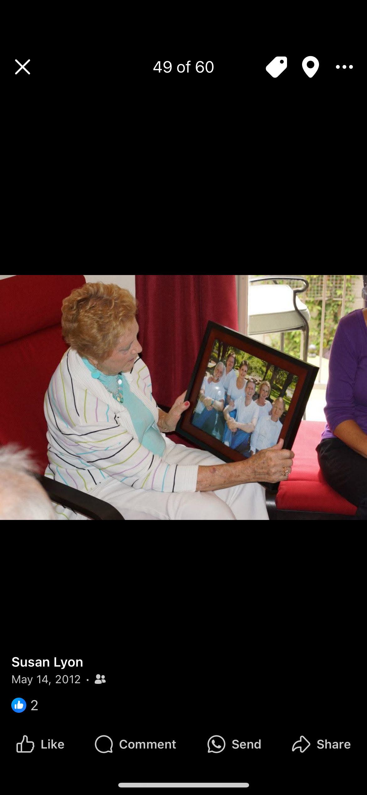 An elderly woman happily shows a family portrait, surrounded by friends in a cozy setting.