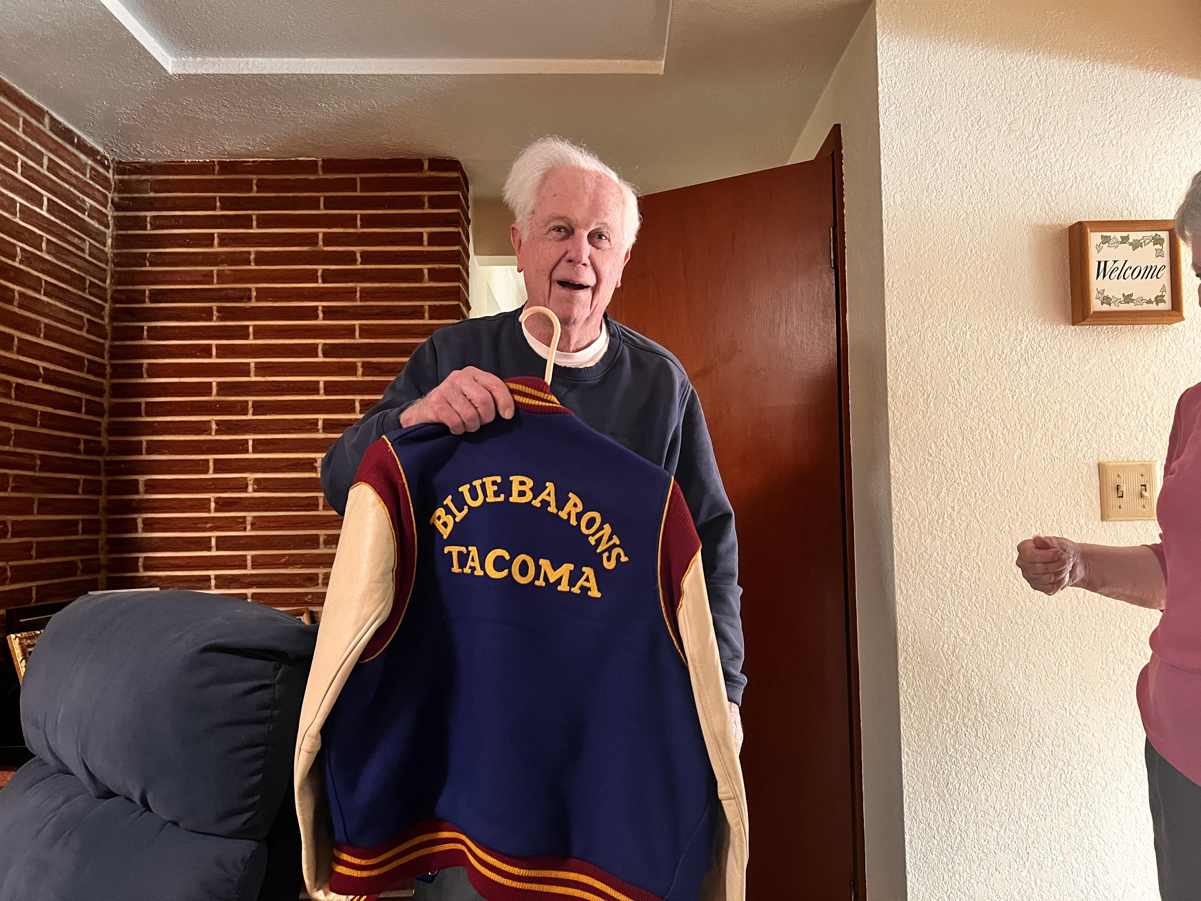 An elderly man smiles while holding a nostalgic varsity jacket during a family gathering.