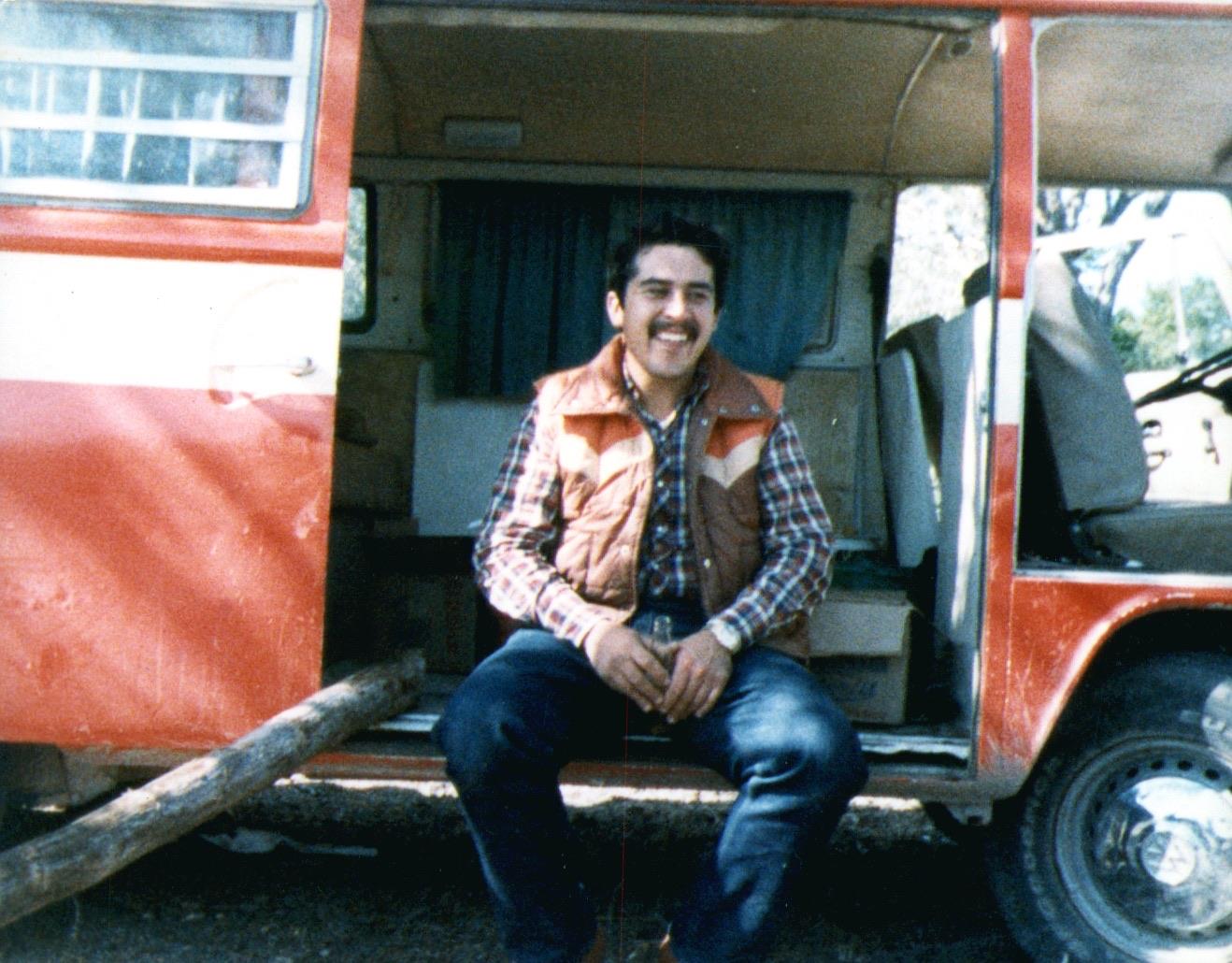A man sits on the steps of a red and white vintage camper van, enjoying the warm sun.