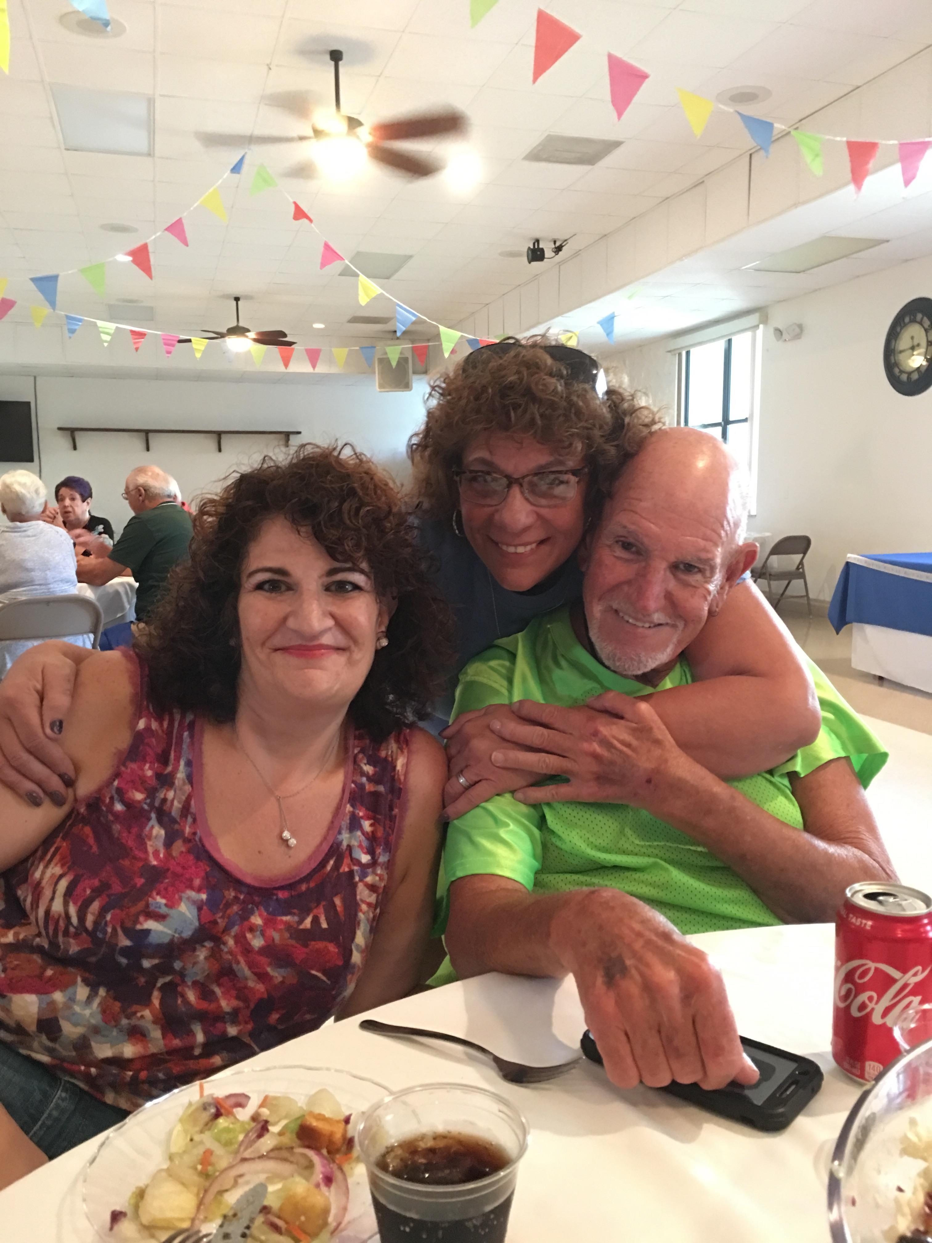 Three friends share a warm embrace while enjoying tasty food at a lively community gathering.