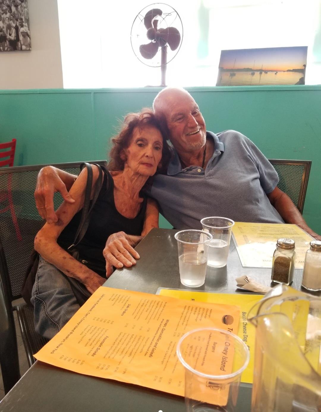 Two elderly individuals share a warm moment at a table filled with menus and drinks.