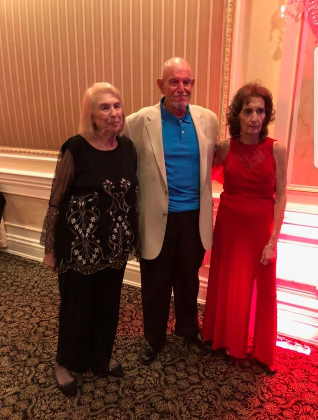Three elderly friends stand together in a festive setting, smiling and enjoying the occasion.