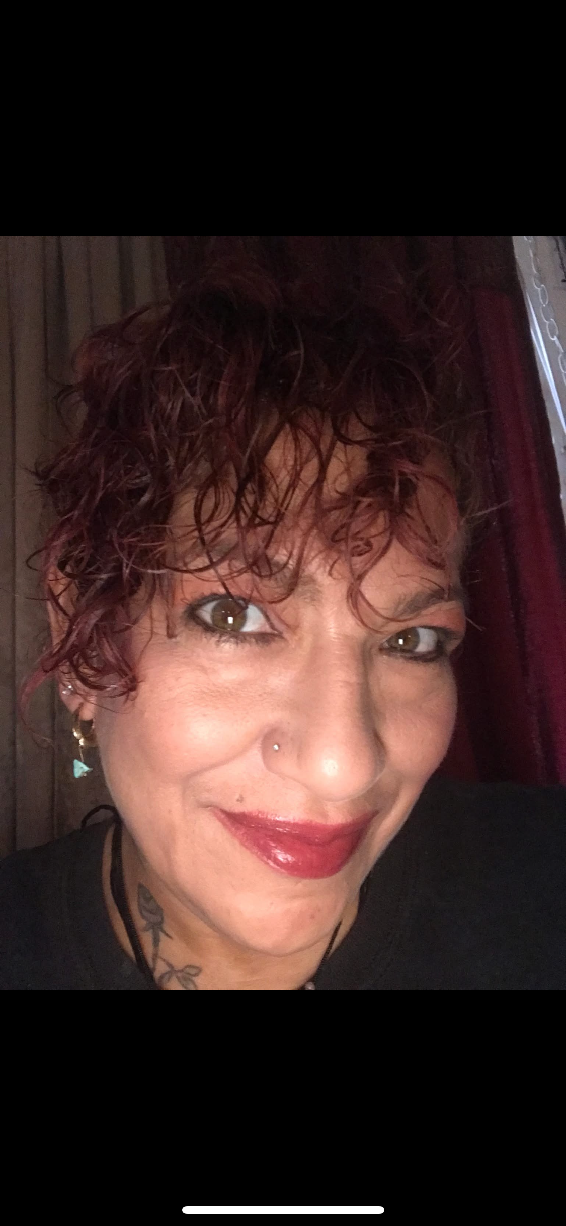 A woman with curly, reddish hair and bold lipstick smiles warmly while sitting indoors.