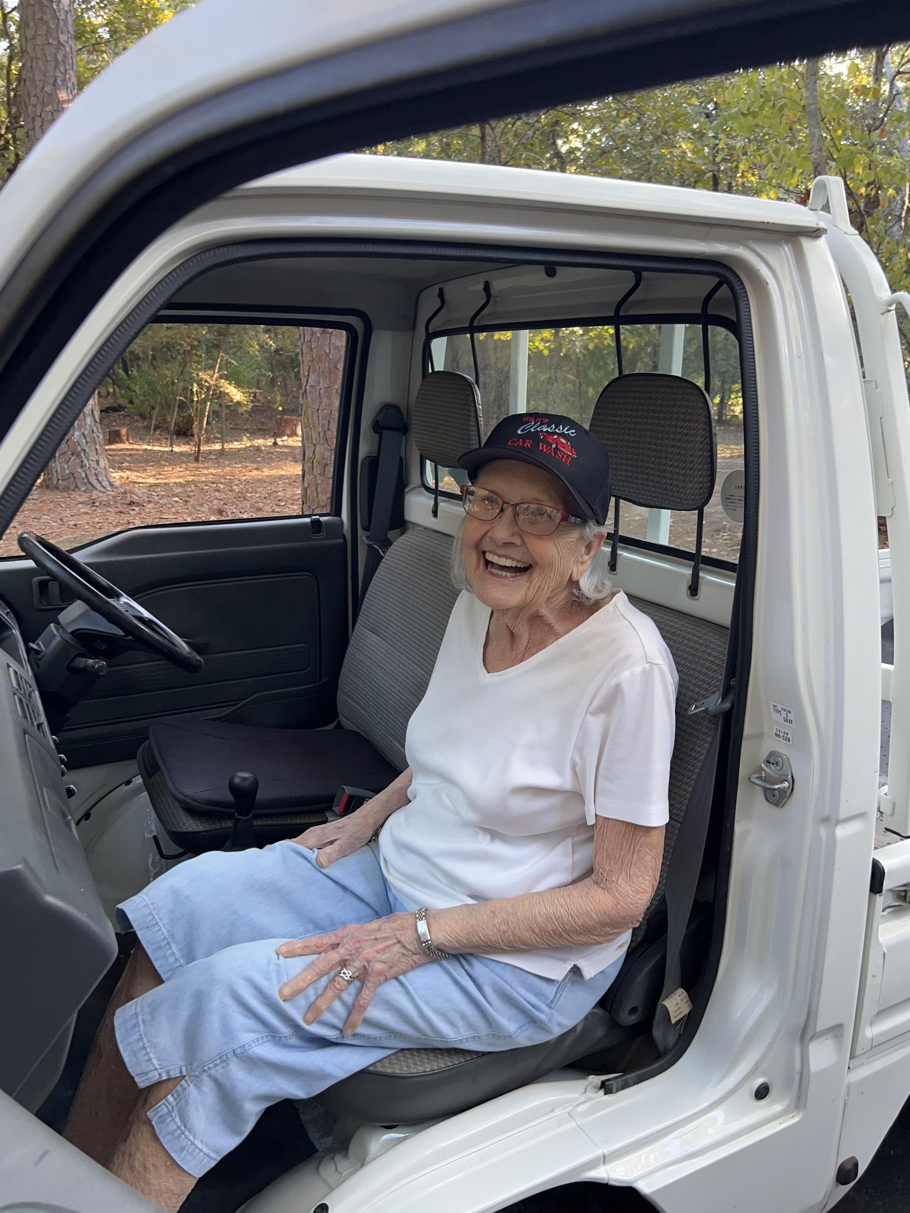 Elderly woman smiles while seated in a parked vehicle, surrounded by trees in bright daylight.