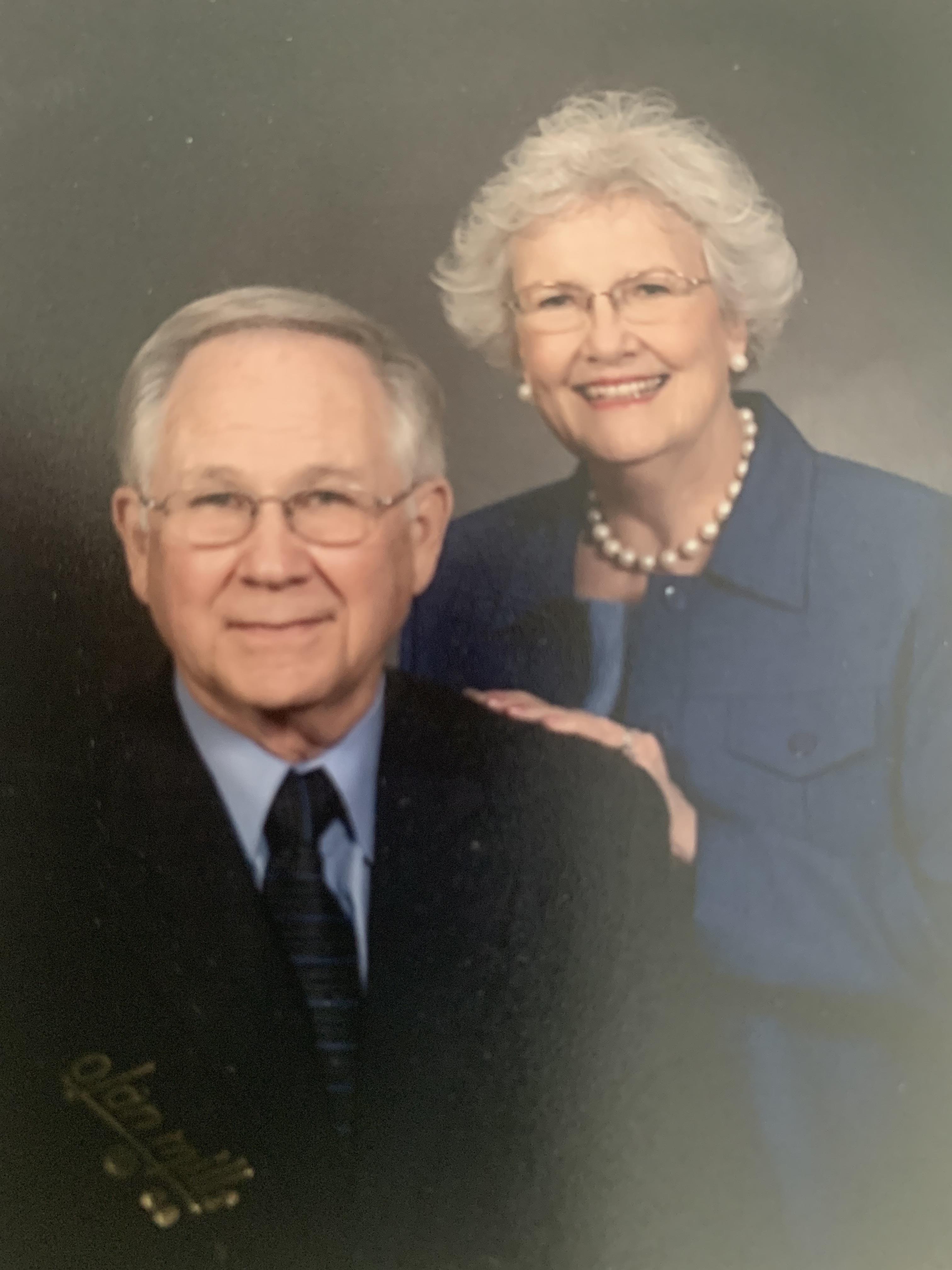 Elderly couple smiles warmly while posing for a formal portrait indoors.