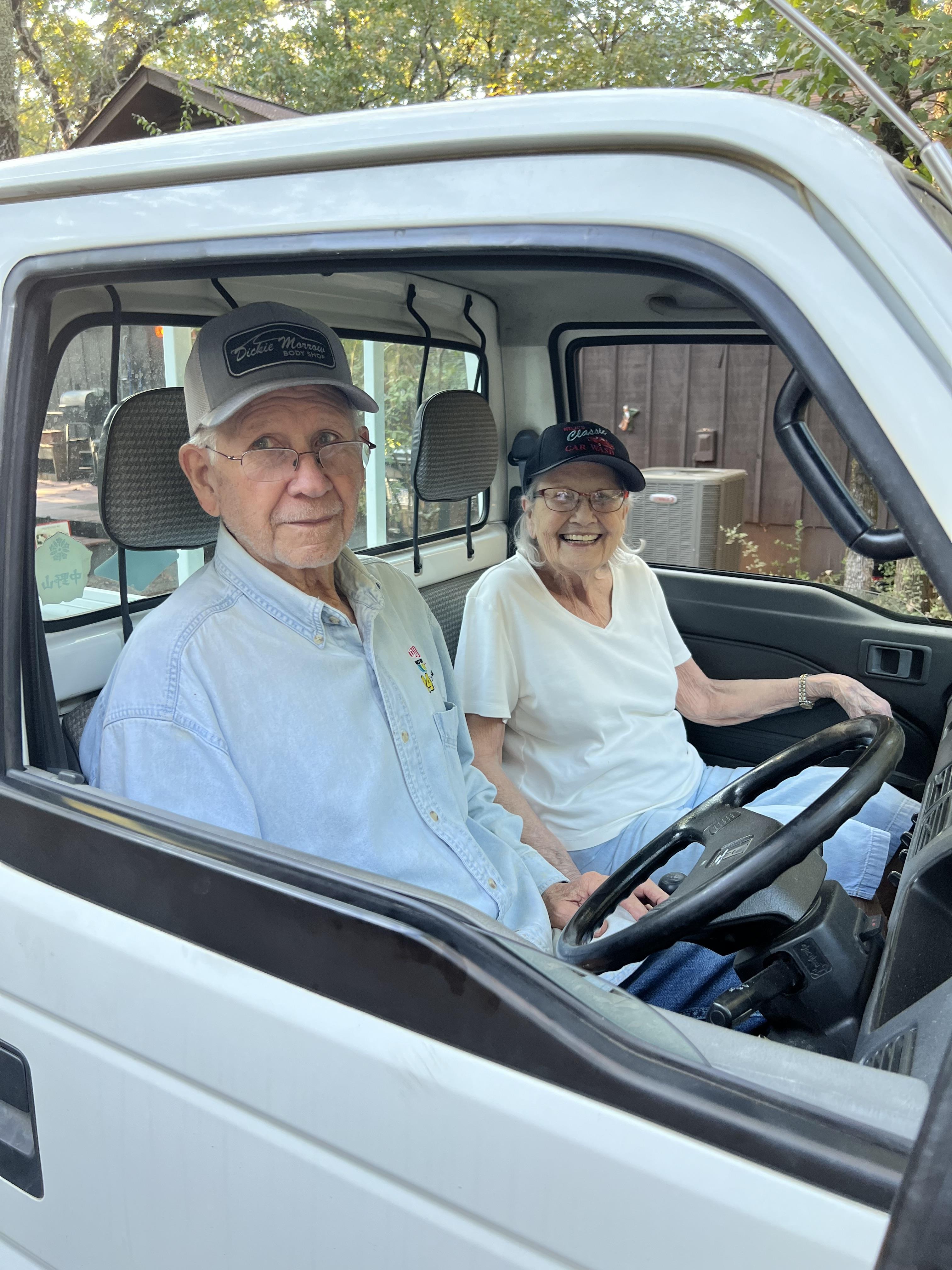An elderly man and woman are sitting comfortably in their pickup truck, smiling happily.