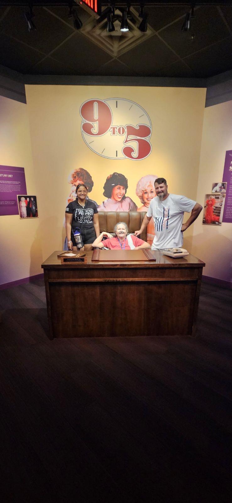 Two visitors pose with an actress in a 9 to 5 themed display at a museum.