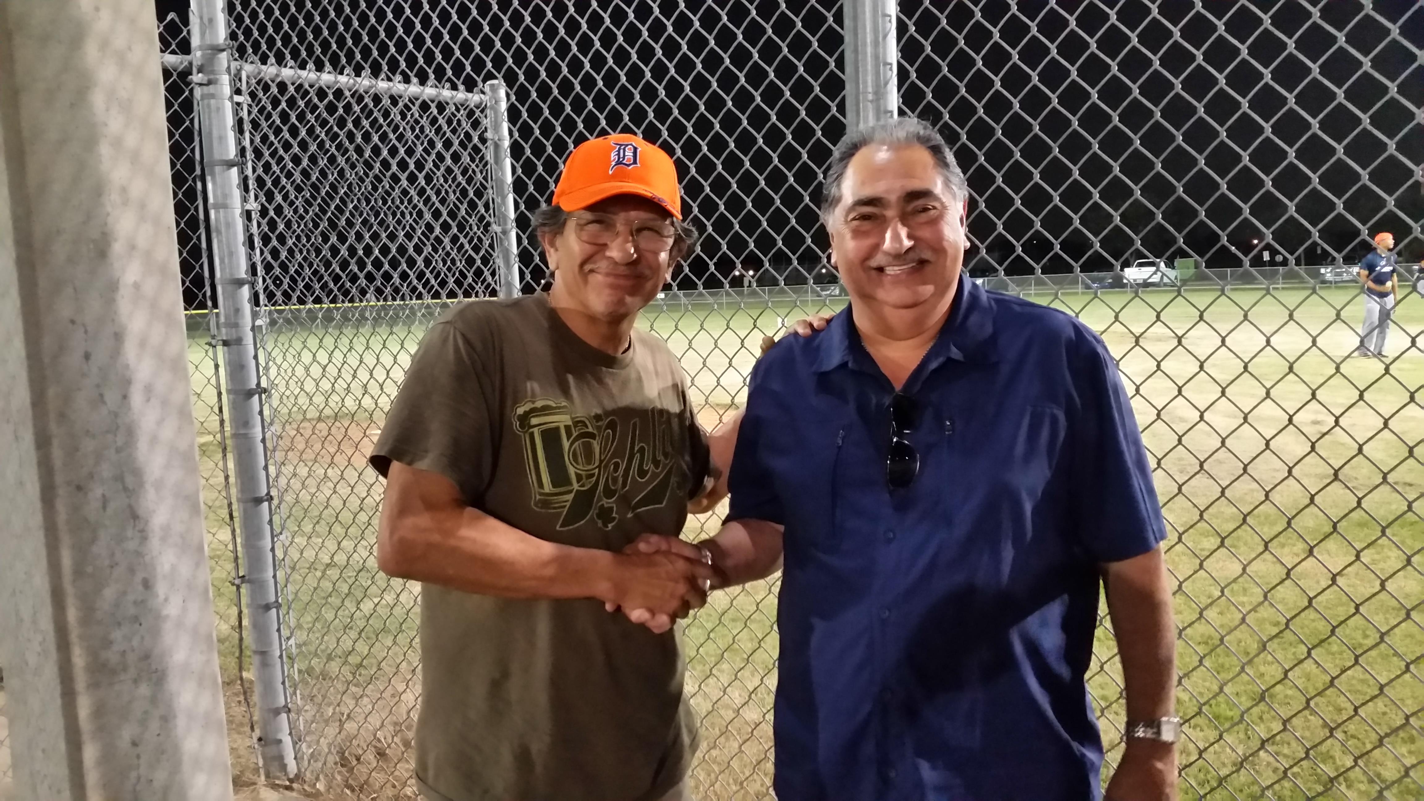 Two men smile and shake hands by the baseball field, creating a friendly vibe.