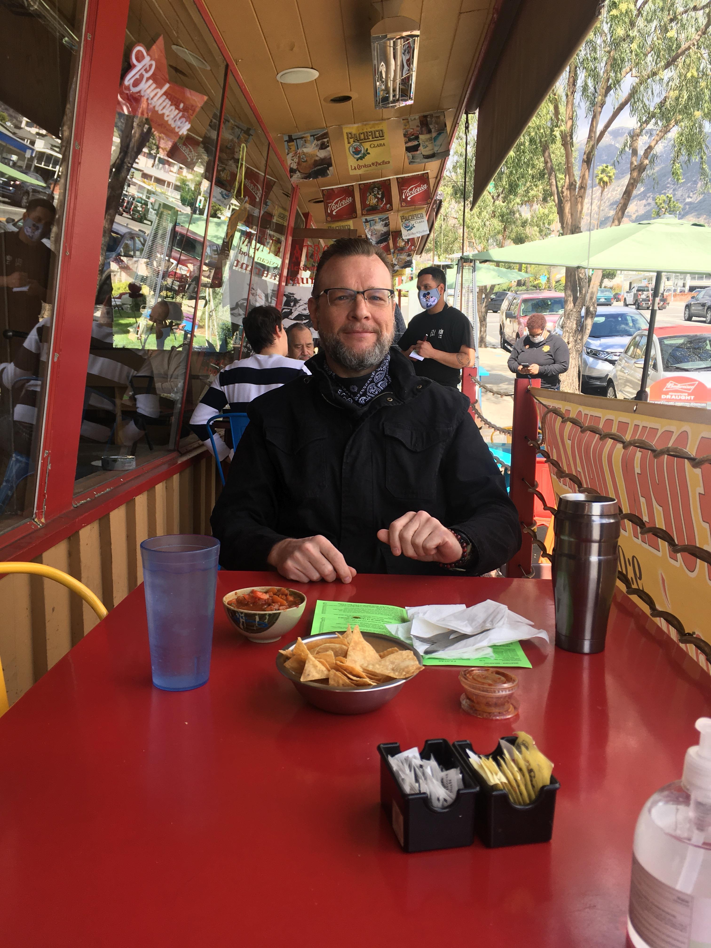 A man sits at a bright red table with snacks, beverages, and a casual atmosphere in the background.