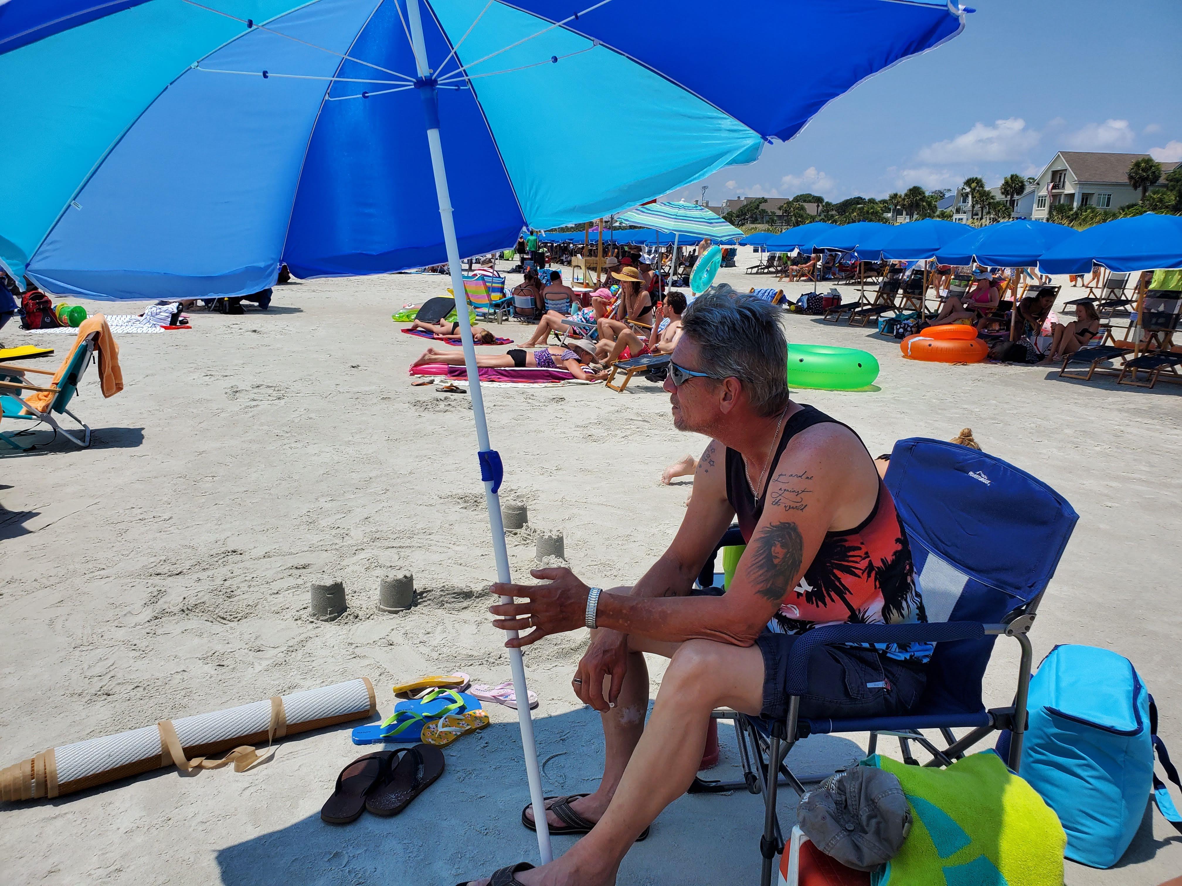 A man sits in a chair under a blue beach umbrella, enjoying the view of beachgoers around him.